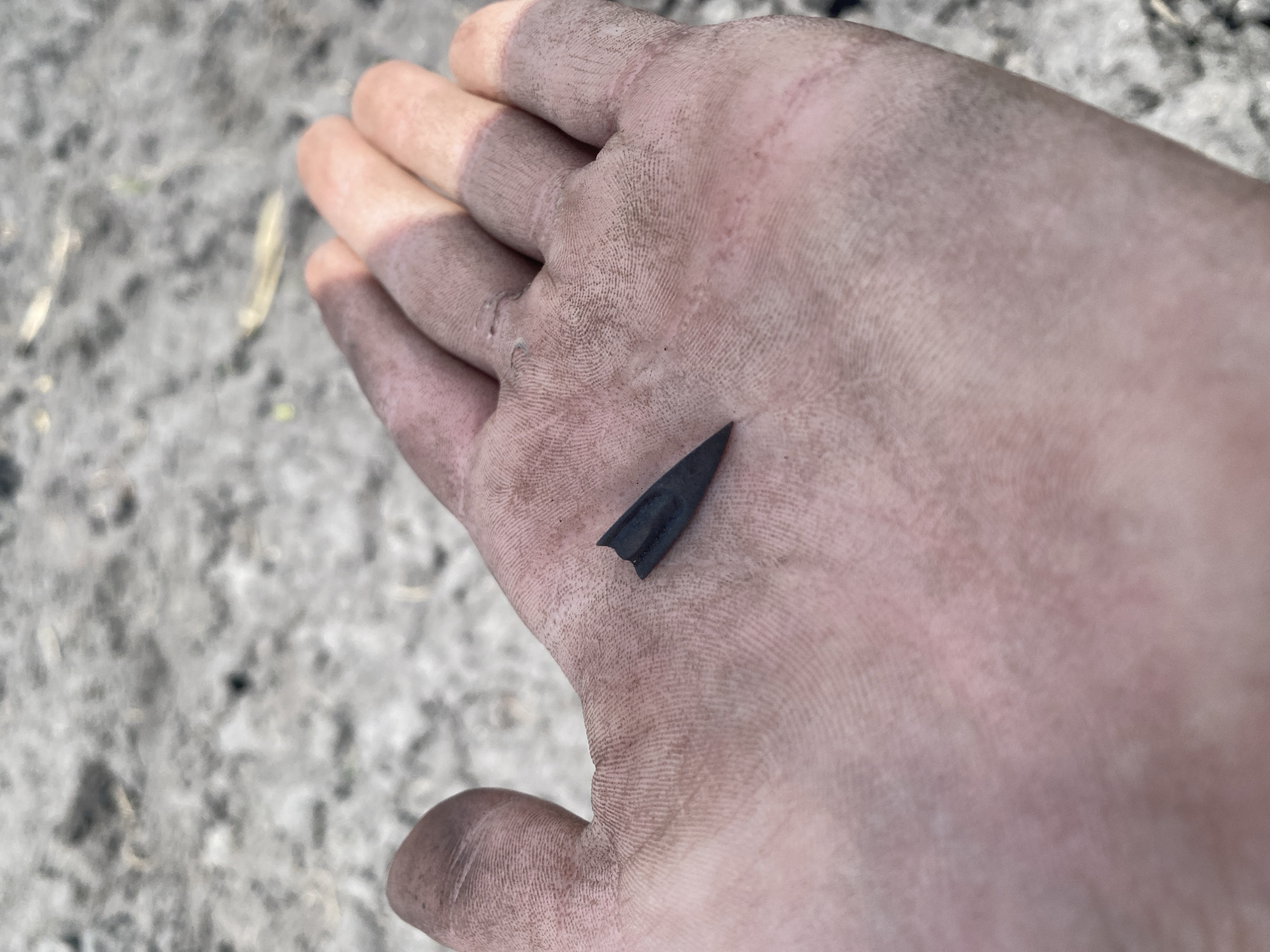 A dirty hand is open with a small, dark-colored, triangular object resting on the palm. The object appears to be an arrowhead. The background consists of grayish-brown textured ground, possibly soil. The hand shows signs of dirt or soil accumulation, particularly along the fingers and knuckles. The photograph was likely taken outdoors during daylight. The location of Matiashivka, Ukraine is not discernible from the visual information provided.