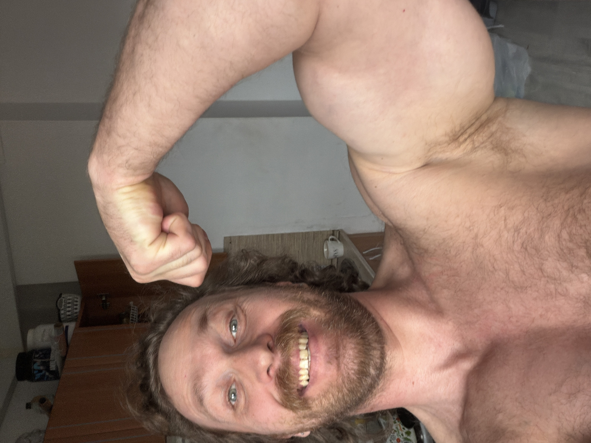 A man with long brown hair and a beard smiles at the camera while flexing his right arm. He is shirtless and appears to be indoors. His facial expression and the flexed arm suggest a display of strength or pride. The background shows a wall and part of furniture. It is impossible to determine the specific location in Saint Petersburg, Russia, based on the image alone.
