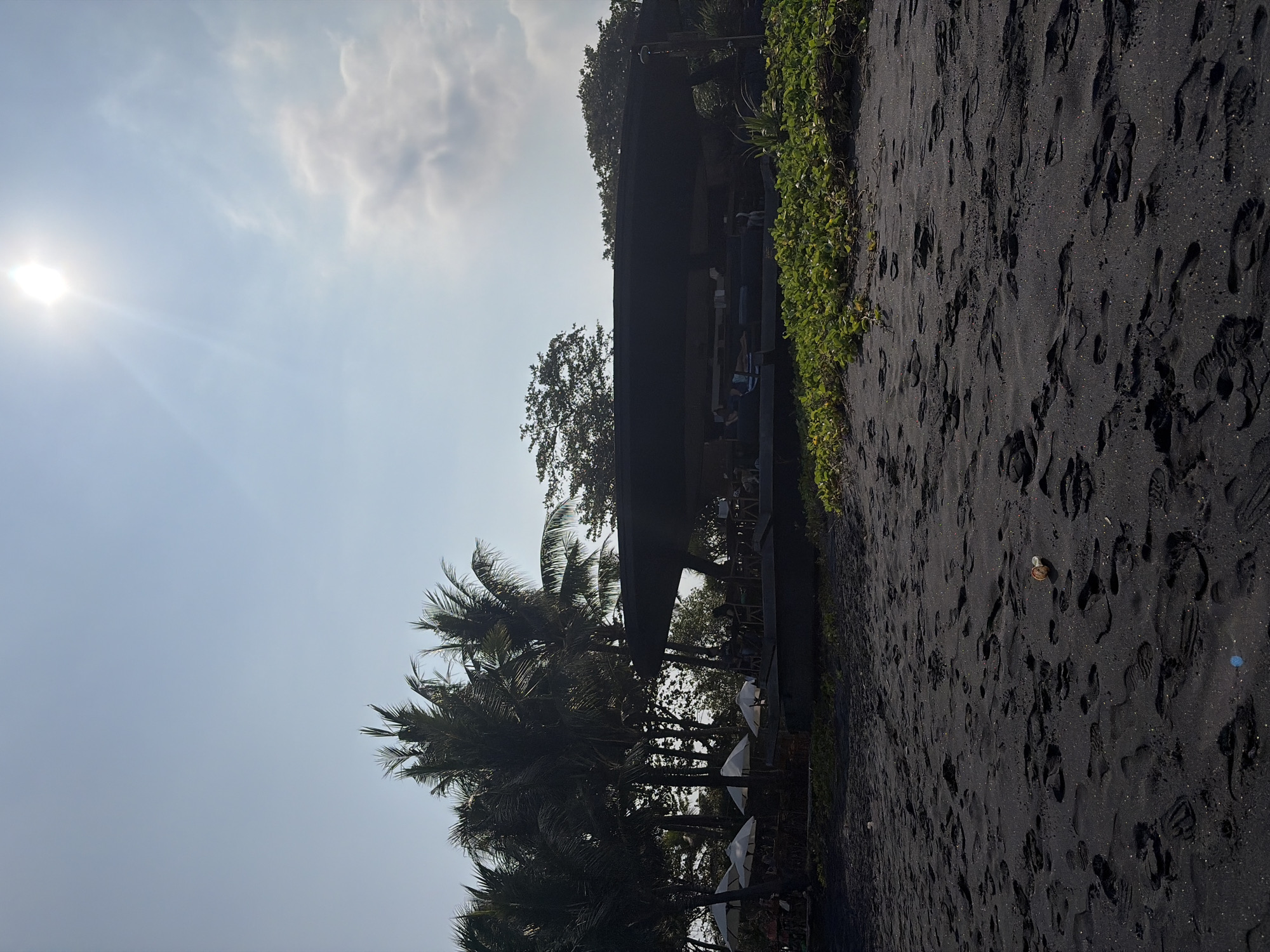 The image depicts a beach scene in Blahbatuh, Indonesia. The foreground is dominated by dark, black sand covered in numerous footprints. A few pieces of debris are scattered on the sand, including what appears to be a small plastic bottle cap. In the mid-ground, there is a lush green patch of vegetation bordering the sand. Behind the vegetation, a low-lying, dark-colored structure with a wide, overhanging roof is visible. This structure appears to be an open-air establishment with seating and tables. Several white umbrellas are set up in front of the structure, providing shade. Palm trees and other foliage are situated behind the umbrellas and the structure, with some trees reaching towards the sky. The sky is a pale blue with a bright sun visible in the upper right quadrant, casting a glare and illuminating some wispy clouds. Several figures are seated at tables or moving around within the shaded area of the structure. The overall impression is a relaxed, seaside location.