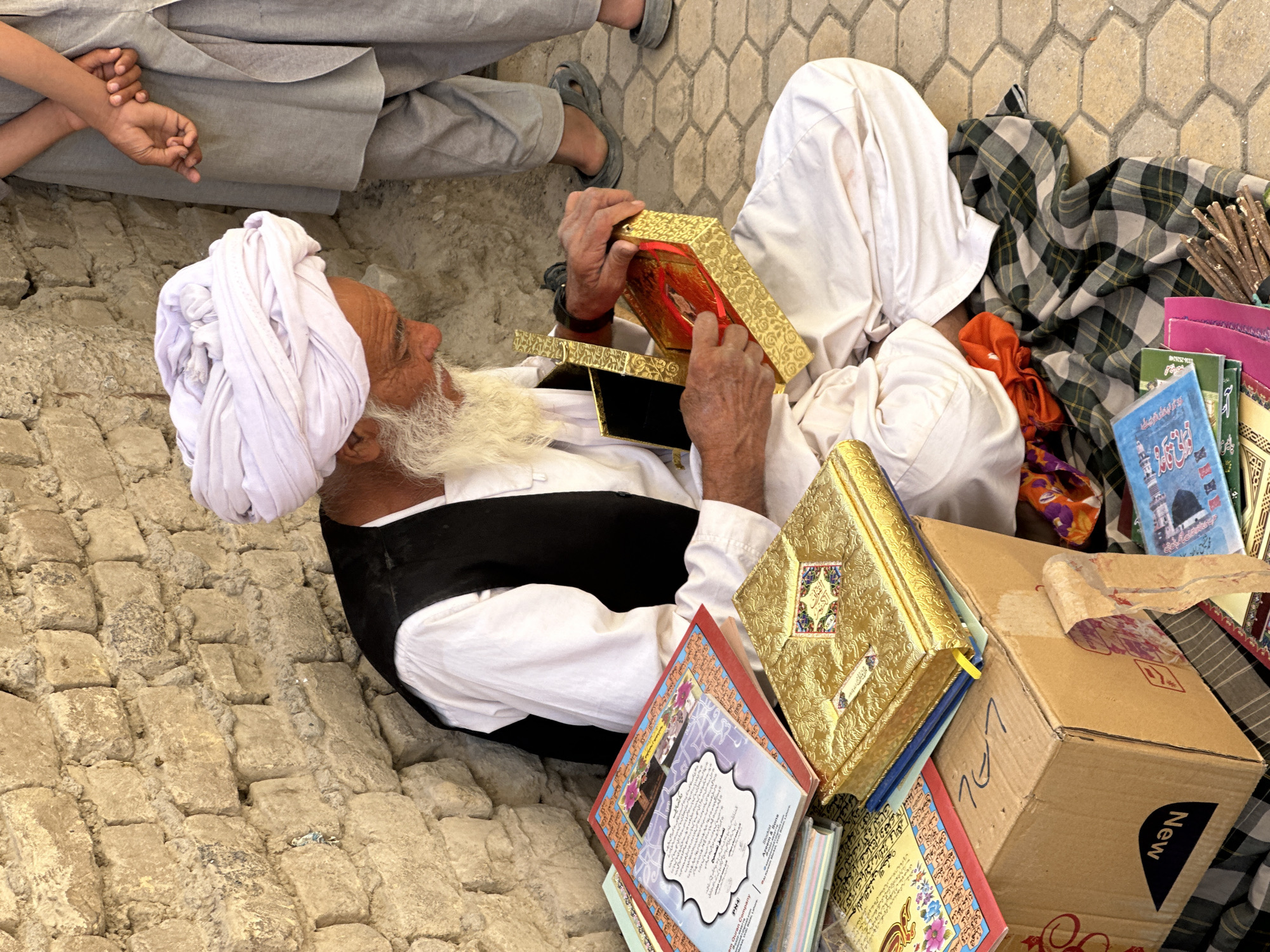 In the city of Kanozai, Pakistan, an elderly man with a white beard and a large white turban is seated on the ground next to a brick wall. He is wearing a white shirt and a black vest. In front of him, a collection of books and boxes are displayed on the ground. The man is holding a small, ornate golden box open, revealing a red item inside, and appears to be examining it. The ground is paved with hexagonal stones. Another person, partially visible, stands to the right, wearing a grey traditional outfit. The scene depicts a vendor or collector of books and religious items, possibly engaged in a transaction or presentation.
