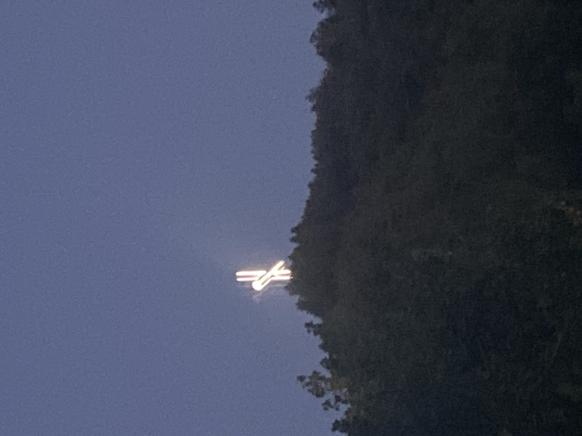 The image shows a nighttime or twilight view of a hill in Blagoevgrad, Bulgaria. Atop the hill, a large illuminated cross is visible against the dark blue sky. The cross appears to be made of light tubes or bulbs, giving it a glowing effect. The hill itself is covered with dark green trees, which are silhouetted against the sky. The overall scene is a serene and somewhat dramatic depiction of a religious landmark in its natural setting.