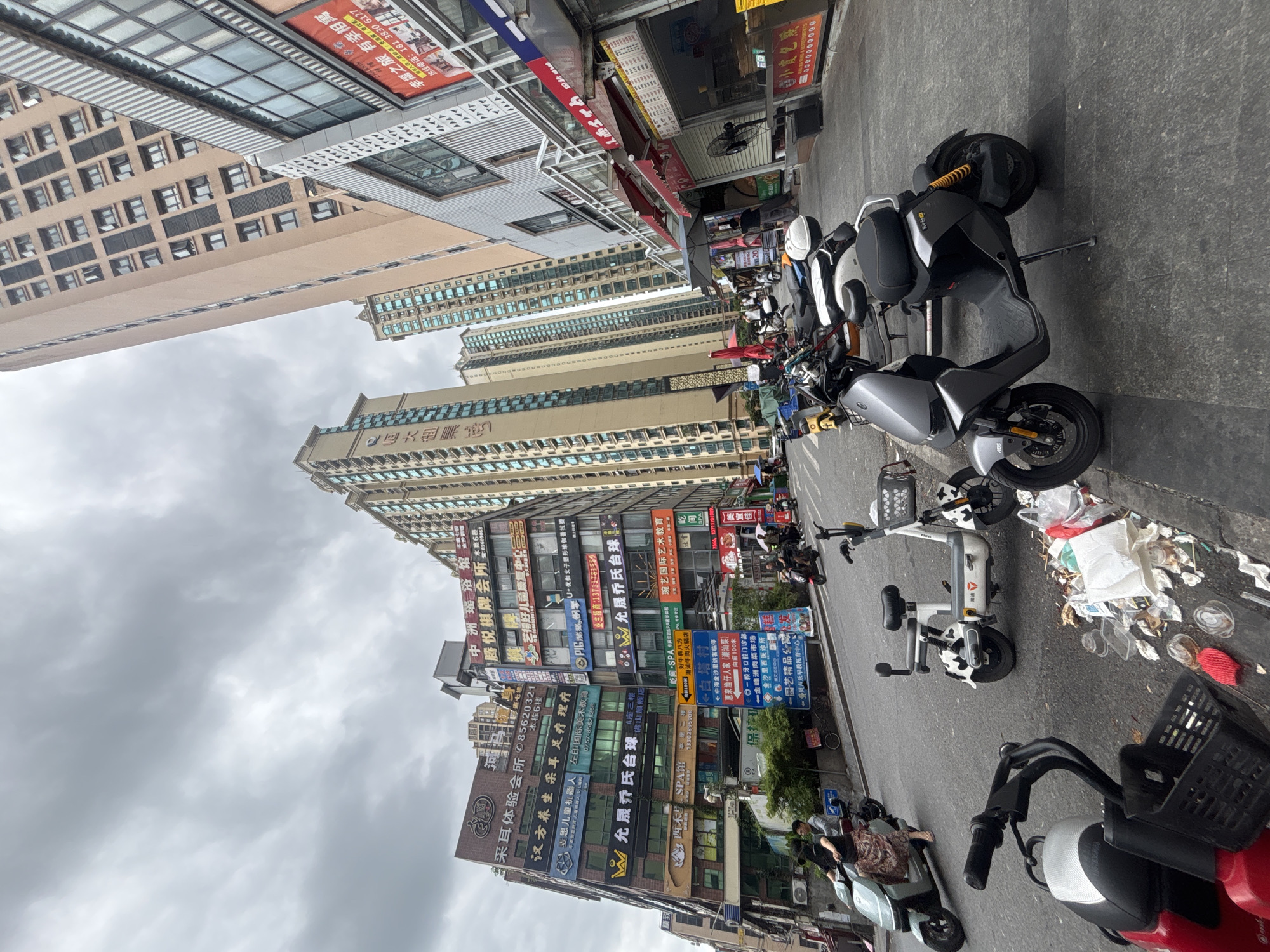 The image depicts a street scene in Nanhai District, China. The perspective is low, looking up at tall, modern buildings lining a street. The sky is overcast with gray clouds. In the foreground, several electric scooters and motorcycles are parked along the side of the road. Some scooters are a lighter color with cow-like patterns, while others are darker gray and red. There is a considerable amount of litter on the curb and street.
Several people are visible, some riding scooters. One person on a scooter in the mid-ground is holding an umbrella. Others are walking on the sidewalk. The buildings are covered with numerous signs, many of which appear to be in Chinese characters, indicating commercial establishments. The architecture is a mix of older, lower buildings with many signs and newer, tall residential or commercial towers. The street appears to be paved, and there are some trees and bushes visible near the buildings on the left. The overall impression is a busy urban environment.