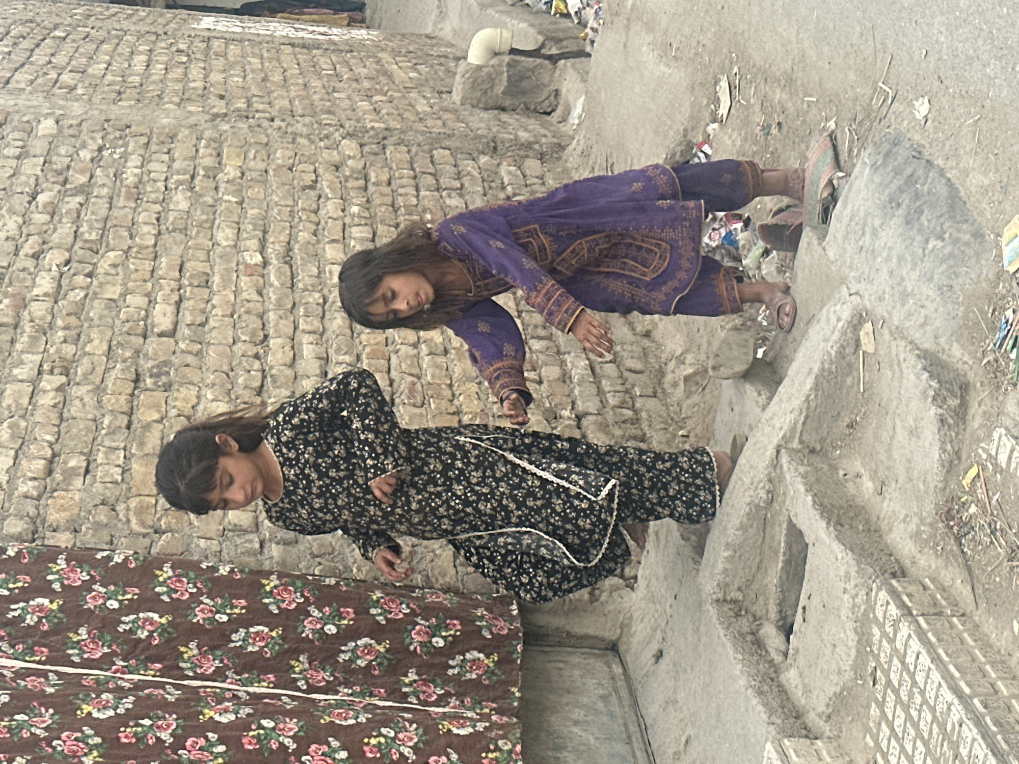 Two girls are in an urban environment, likely Quetta City Tehsil, Pakistan. One girl wears a dark floral dress, and the other wears a purple dress with gold embroidery. They stand near a brick wall and what appears to be a concrete structure, possibly a drainage system. A floral-patterned mat or fabric sits nearby. The surrounding ground shows evidence of trash and debris.
