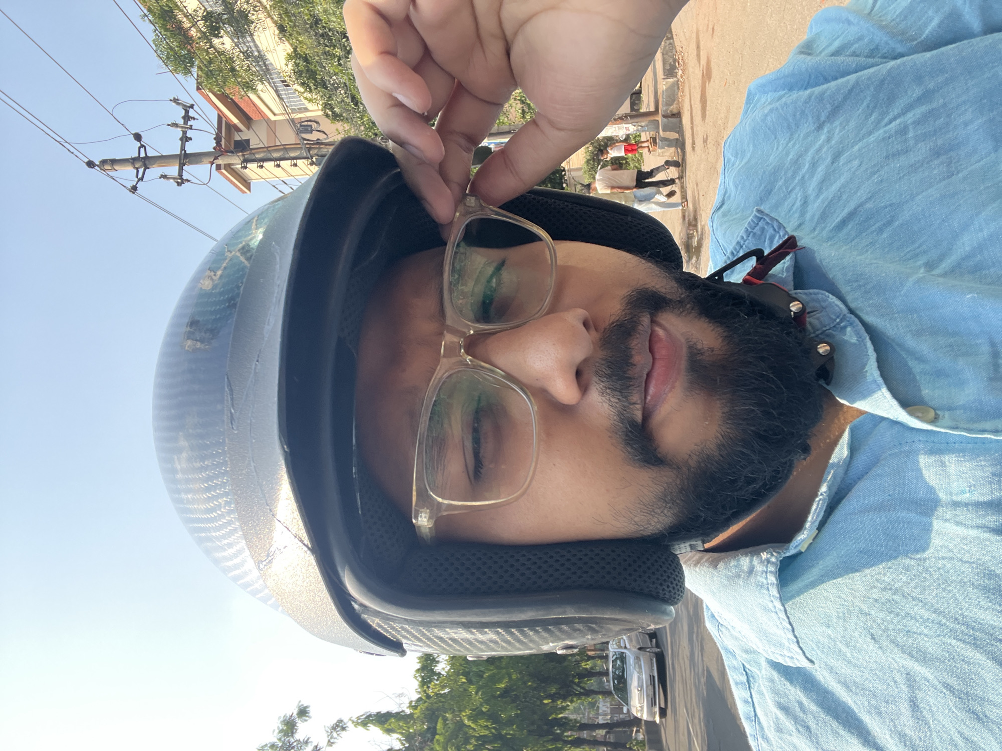 The image shows a person wearing a helmet and glasses, adjusting the glasses with their hand. They are wearing a light blue shirt. The background includes a street with trees, buildings, and utility poles. In the distance, people are walking. The location is possibly in the city of Dhaka, Bangladesh.