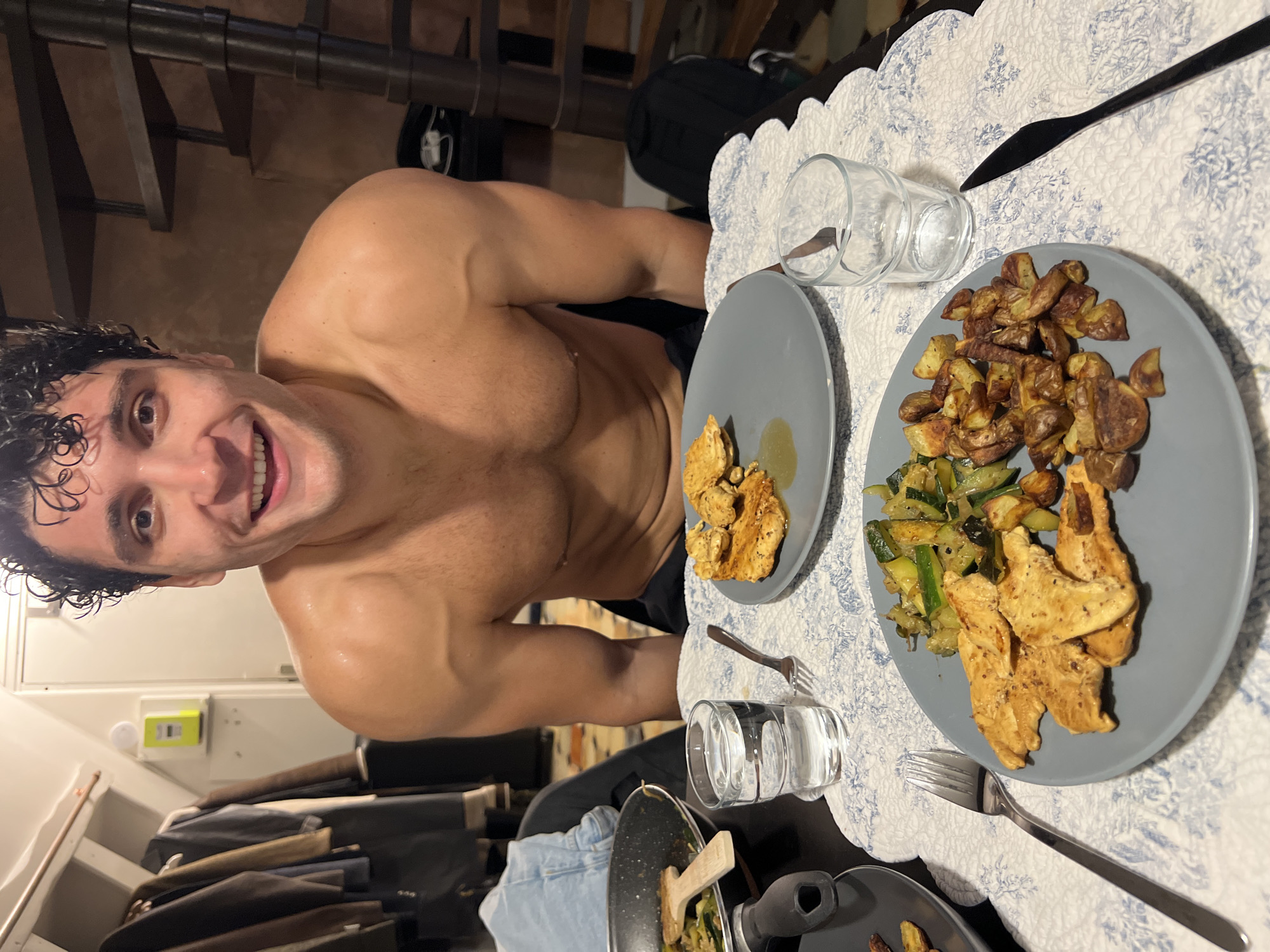 A shirtless man with wet, curly dark hair smiles at the camera while sitting at a table with two plates of food. The plates contain grilled chicken, roasted potatoes, and sautéed zucchini. There are two glasses of water and silverware on the table, which is covered by a white and blue patterned tablecloth. In the background, there are coats hanging on a rack, a spiral staircase, and a yellow electrical box on the wall. The overall setting appears to be a residential interior, possibly in Paris, France, given the presence of a spiral staircase and the general aesthetic.