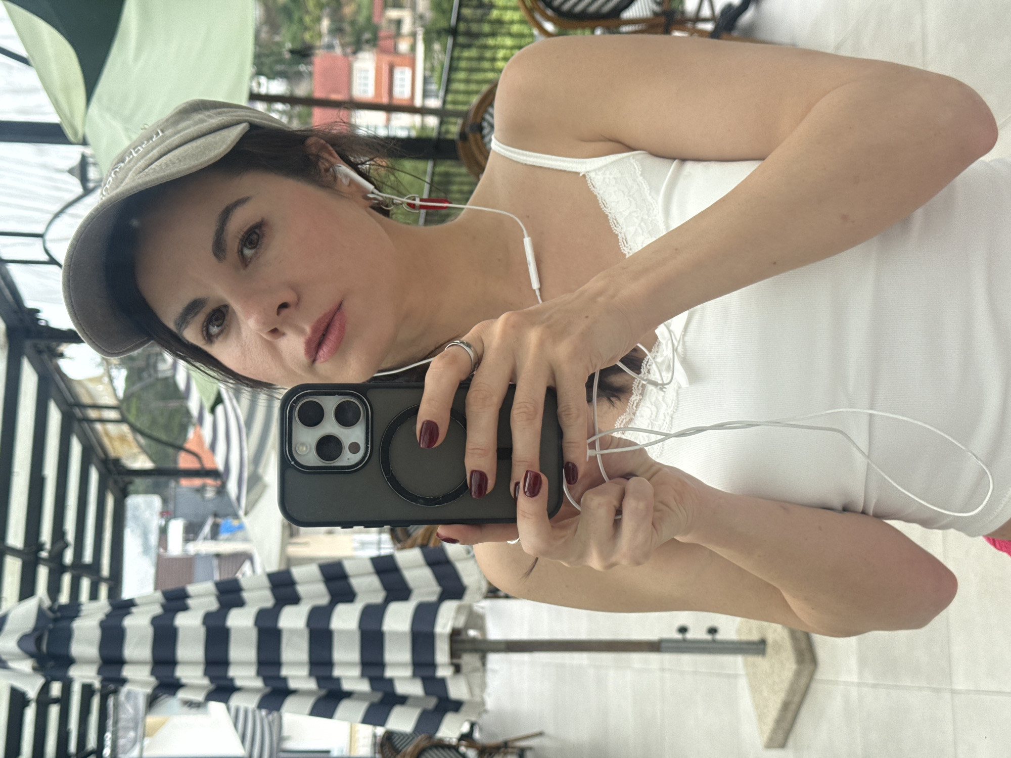 An adult woman wearing a beige baseball cap and a white tank top takes a selfie with a smartphone in a restaurant or cafe with an outdoor seating area in Da Lat, Vietnam. The woman has dark hair and is wearing white earbuds with a red accent. She is also wearing a ring on her left hand. In the background, there are striped umbrellas, tables, chairs, and buildings with red roofs. The overall setting suggests a casual, open-air environment in a city.