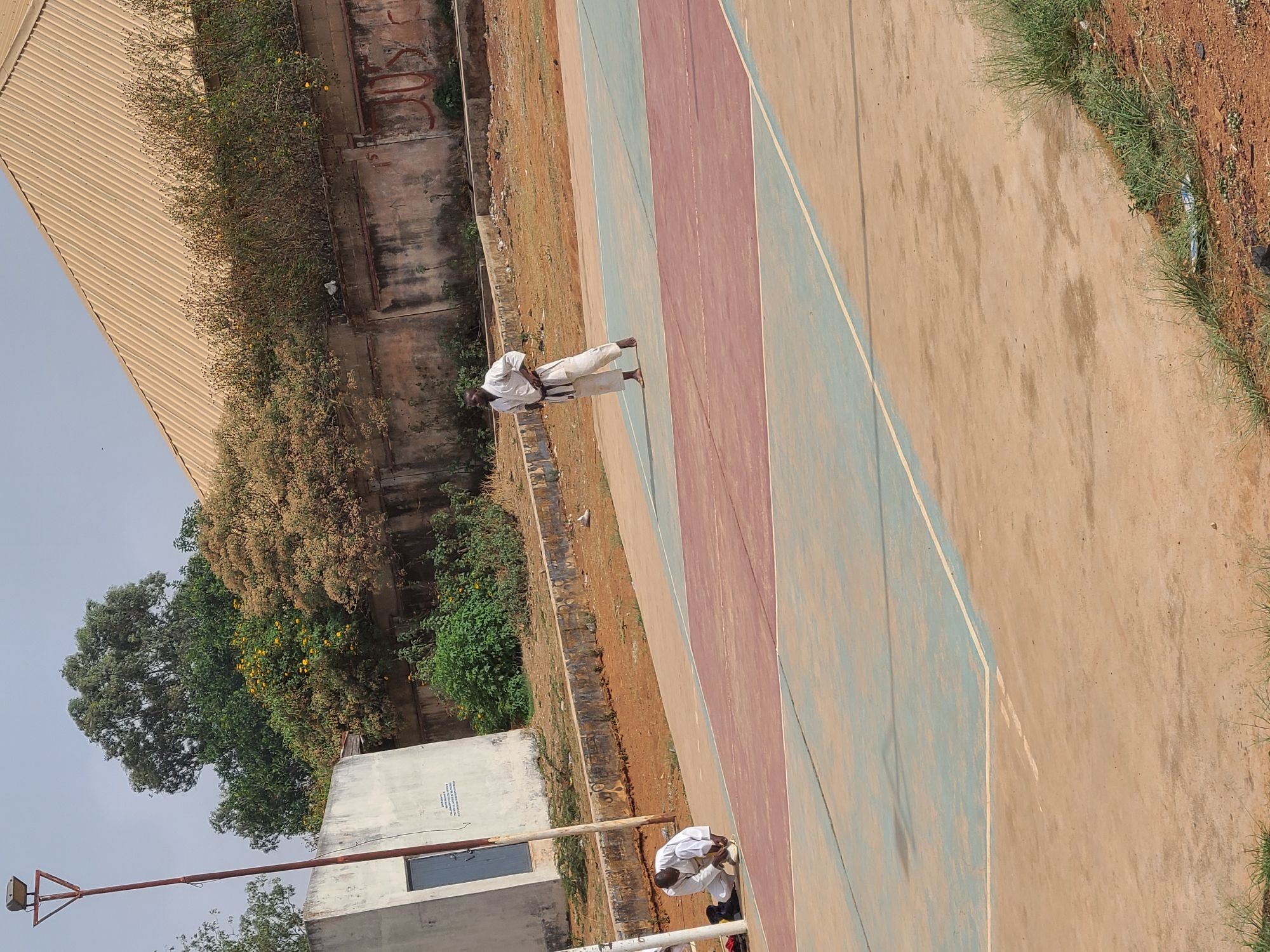 A tilted outdoor shot depicts a building on the left and a painted athletic court on the right. In the middle, two people in white martial arts uniforms are visible. One person stands upright with a black belt, and the other is kneeling. The environment includes trees, bushes, and a building on the left. The location is inferred to be Jos, Nigeria, based on contextual information.