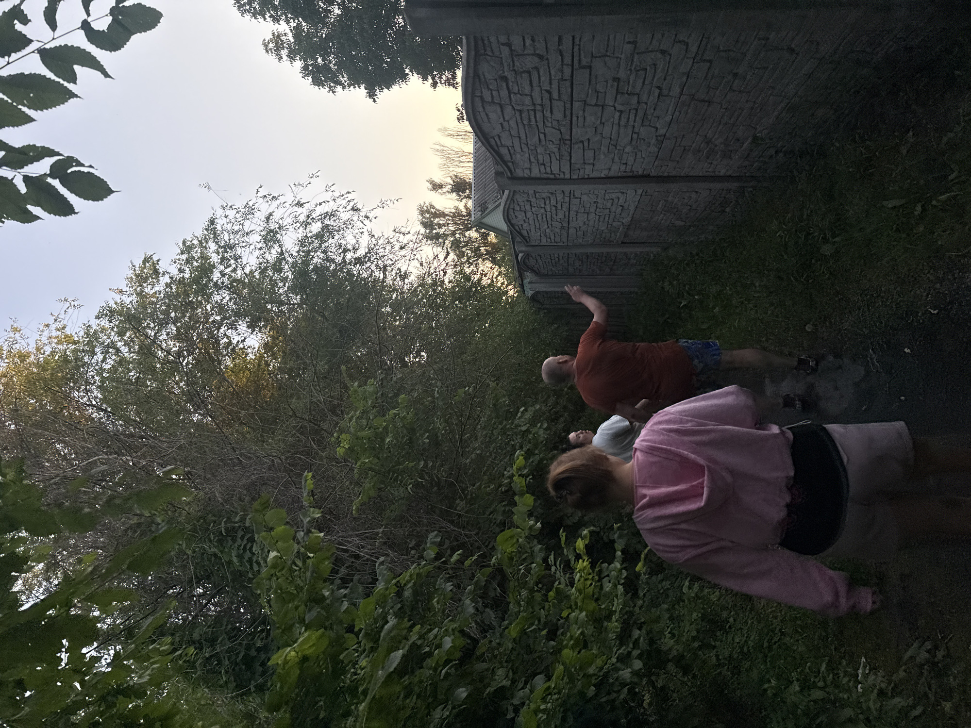 In Obukhivka, Ukraine, three individuals are walking along a path adjacent to a textured concrete fence. The person in the foreground, viewed from behind, wears a pink hooded jacket and light-colored shorts. To their right and slightly ahead, a person in an orange t-shirt and blue patterned shorts is visible, with their right arm extended. Another individual in a white t-shirt is positioned between the first two. The path is bordered by dense green foliage on the left and the concrete fence on the right. The sky above displays a soft twilight glow, with trees visible in the background. The scene captures a moment of casual movement in an outdoor, possibly suburban, environment.