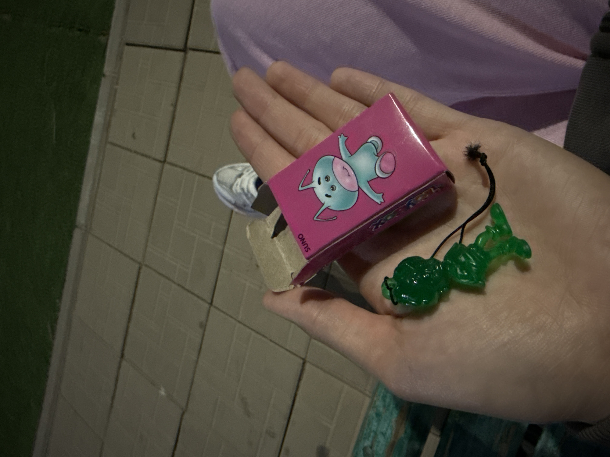 The image shows a close-up of a person's hand holding a small pink box with a cartoonish blue character on it. The box has some text on it, which appears to read "SUNO". Also in the hand are two small, green, plastic figures connected by a black string. One figure resembles a frog or a goblin. The person is wearing a light purple shirt. In the background, a tiled walkway is visible, with a patch of green grass to the left. The lighting suggests it might be evening or a dimly lit area. The person's hand is resting on their lap. The overall scene appears casual and everyday.