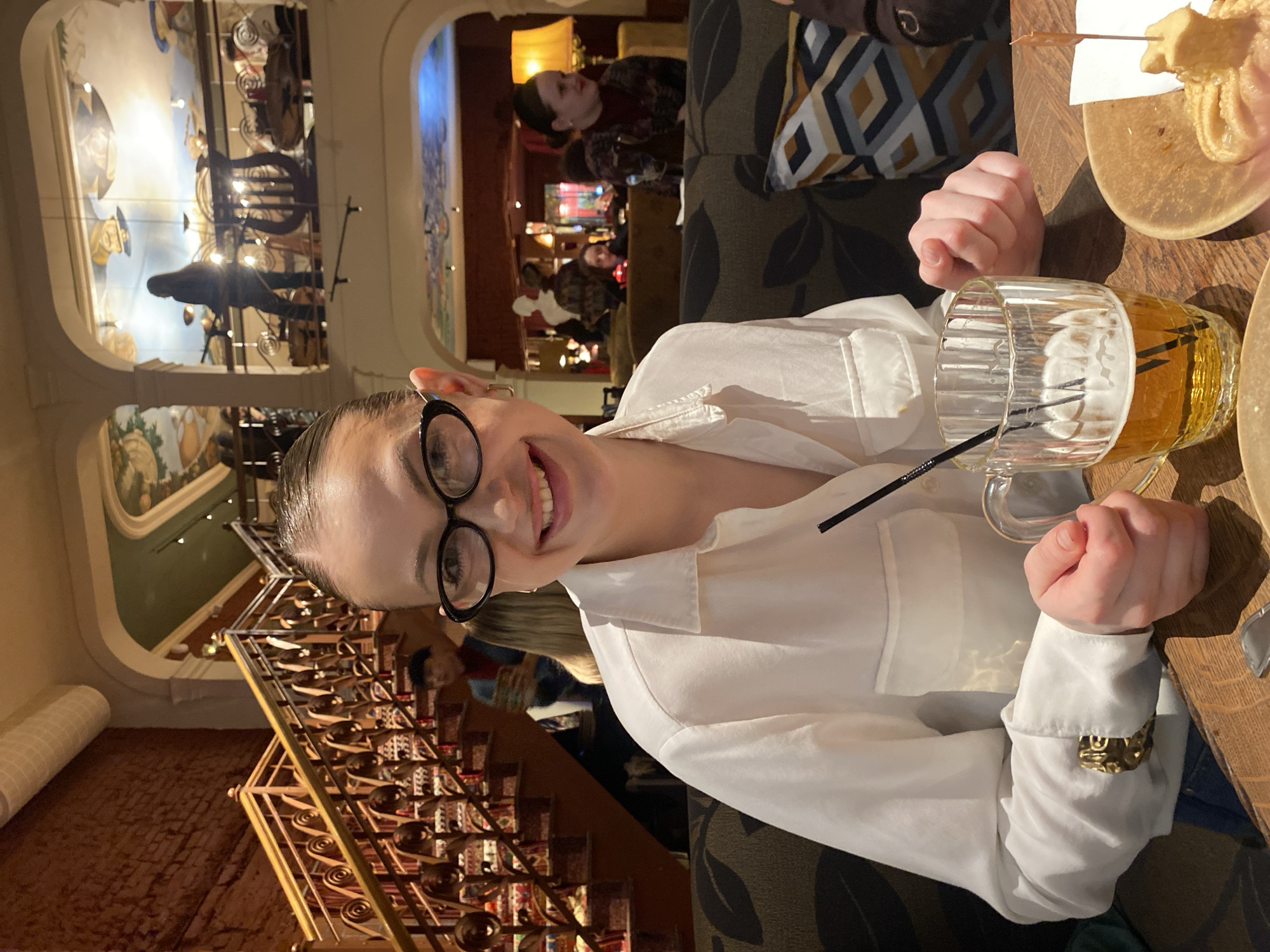 The image captures a woman seated at a table in a restaurant, likely in Kazan, Russia, based on the architectural features visible in the background, including murals and decorative elements. The woman is wearing a white collared shirt and round eyeglasses, and she is smiling towards the camera. On the table in front of her, there is a mug filled with beer and a plate of food, possibly churros. The table is made of wood. The background shows other patrons in the restaurant, along with a grand staircase and decorative murals on the walls and ceiling. The lighting is warm and atmospheric.