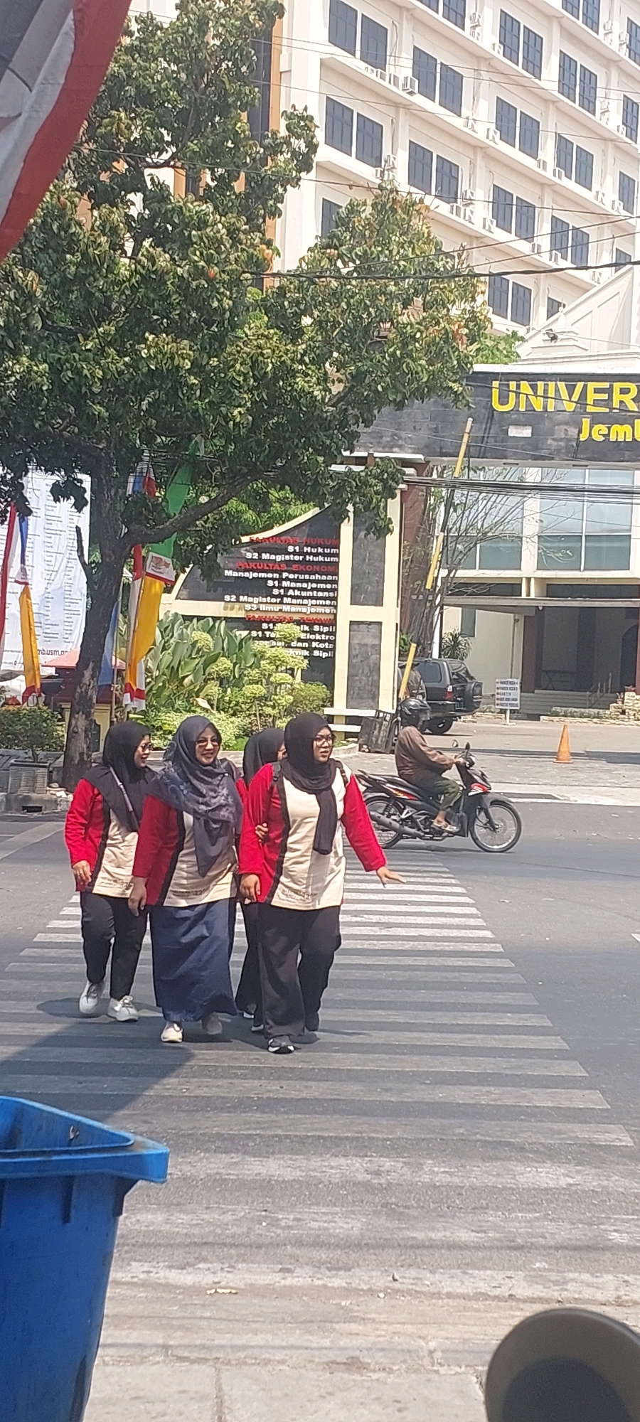 In Semarang, Indonesia, a group of four women wearing headscarves and matching red and beige uniforms are crossing a pedestrian street. They are walking from left to right across a zebra crossing. The woman in the front on the left is wearing white shoes, while the woman next to her is wearing blue jeans and white shoes. The woman in the middle is wearing a long blue skirt and white shoes. The woman on the far right is wearing black pants and black and white shoes. Behind them, a man wearing a brown jacket and helmet is riding a motorcycle. In the background, there is a building with "UNIVERSITAS JEMBER" written on it, suggesting the location is a university. The building is modern with multiple floors and windows. There is also a green tree on the left side of the image, and a blue trash can in the foreground on the left. The scene appears to be during the daytime, with bright sunlight.