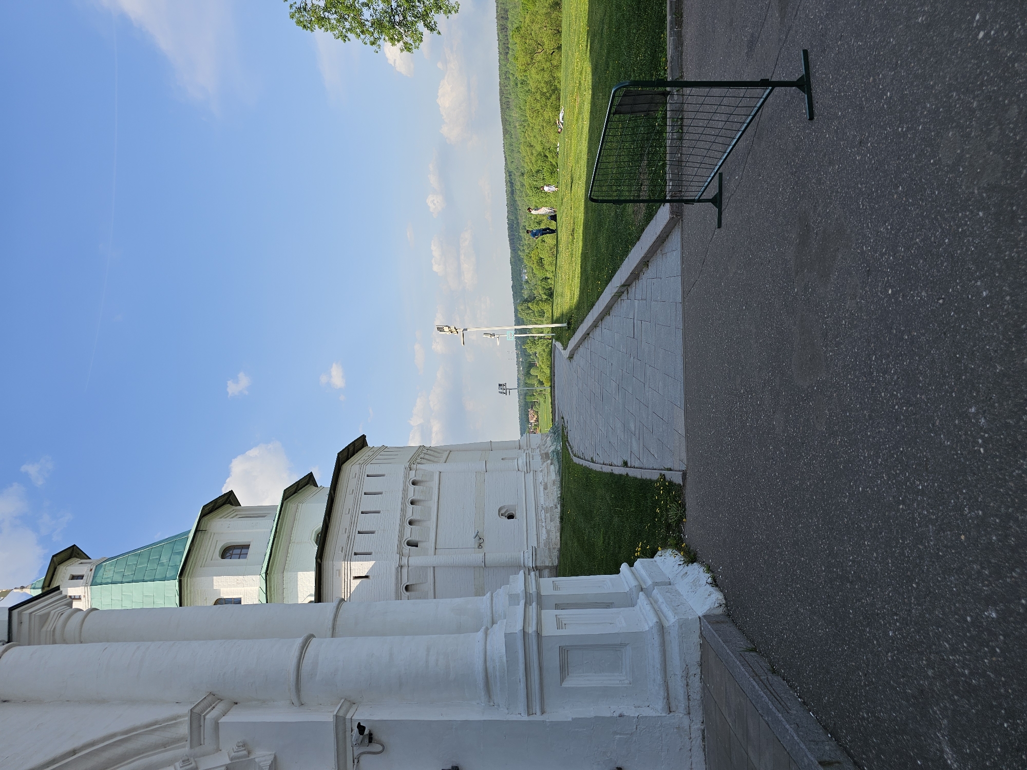 The image depicts a section of the New Jerusalem Monastery in Istra, Russia, taken on a sunny day. The monastery's white walls and green roofs contrast with the blue sky. A walkway with light-colored pavers borders the monastery wall, leading towards a green field. Three people are visible in the distance within the field. A dark asphalt road runs along the right edge of the frame, separated from the walkway by a small section of grass and a green metal fence.