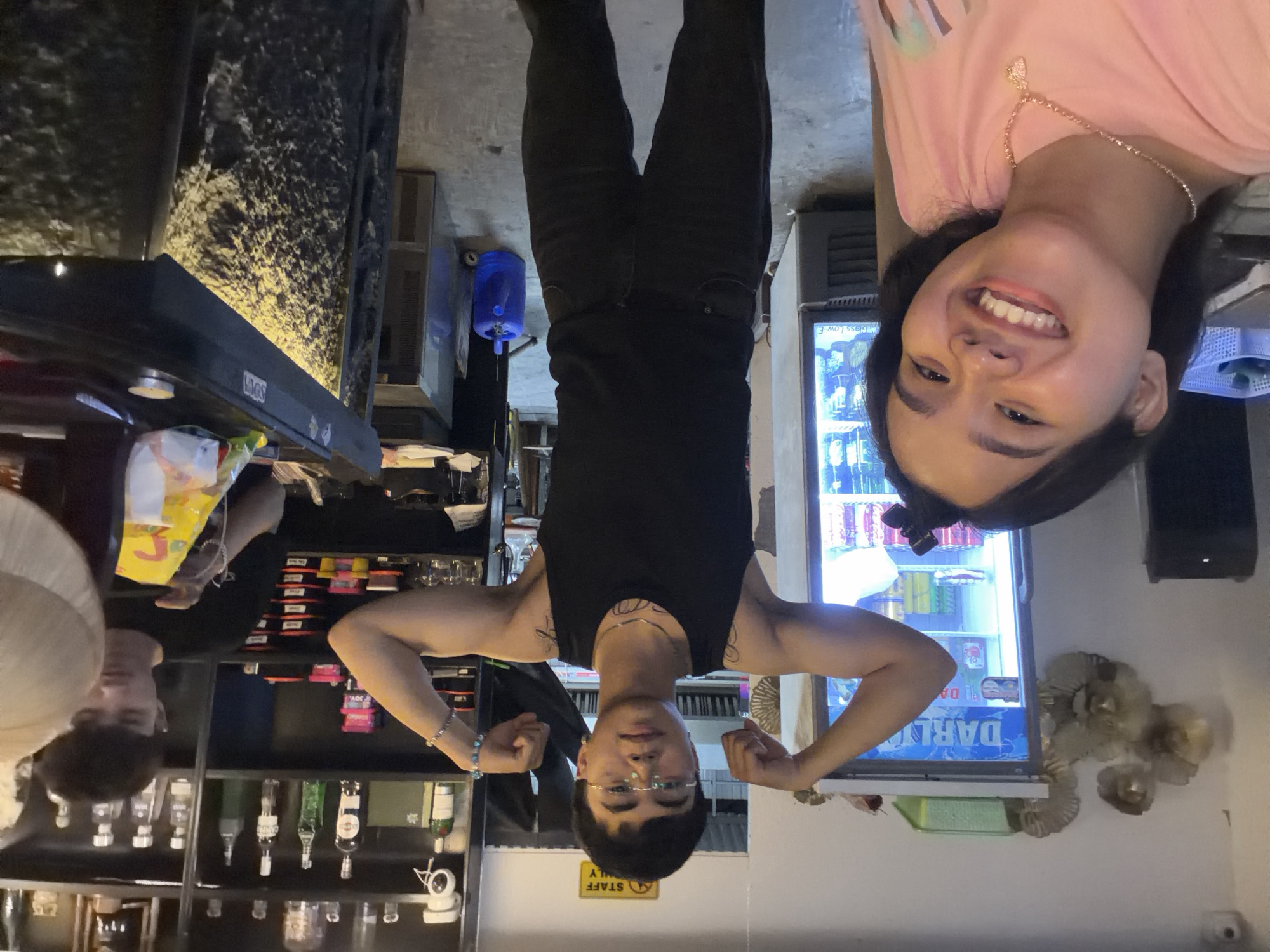 A snapshot taken inside what seems to be a bar setting. Three people are featured. A woman in a pink top smiles at the camera. A man in a black tank top and pants flexes his arms. A man wearing a gray shirt stands on the left side of the bar. On the bar shelves are assorted bottles of liquor and drinking glasses. A cooler is stocked with beverages. The words "DARLA" are printed on the cooler. There is a "STAFF ONLY" sign on the wall. This photo was taken in Đà Nẵng, Vietnam.