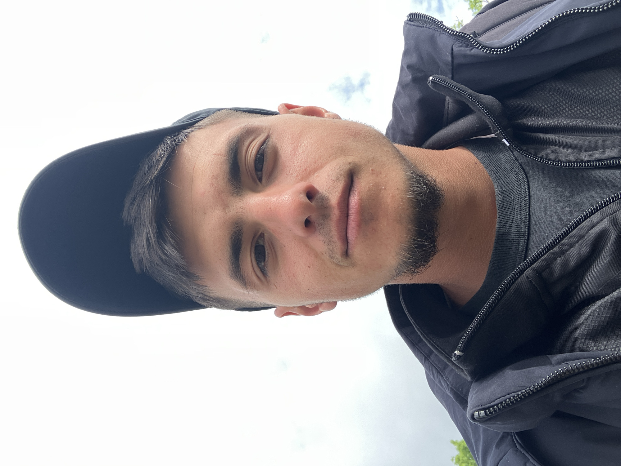 A close-up selfie of a young man is depicted in this image. The man is fair-skinned with dark hair and a stubble beard. He is wearing a dark-colored baseball cap and a black jacket over a dark t-shirt. The background is a cloudy sky. The location cannot be determined as Krastevich, Bulgaria.