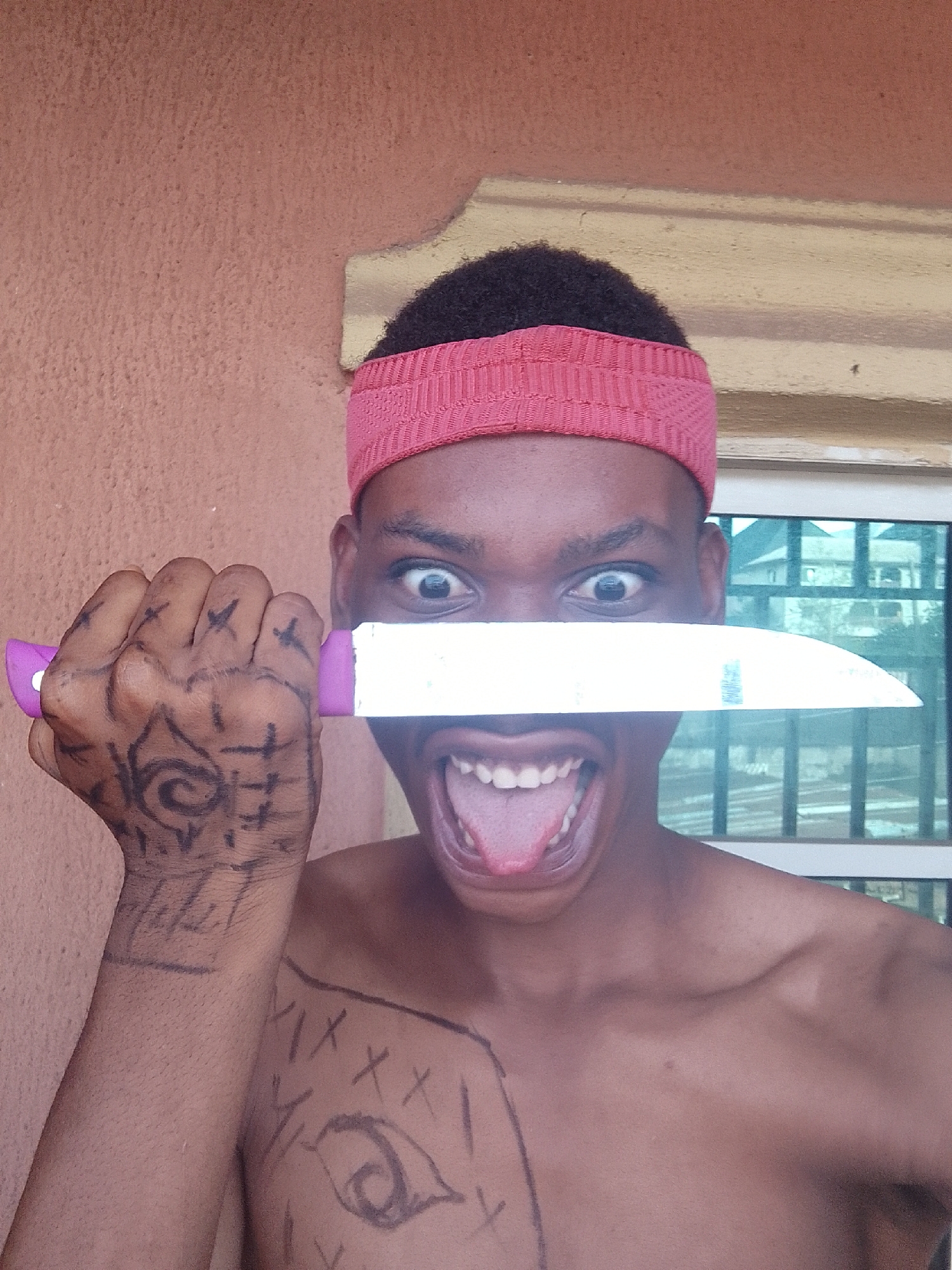 A person with dark skin is holding a knife in front of their face. They are wearing a pink headband and have drawn markings on their body with what appears to be a marker. The person's mouth is open, tongue extended, and eyes are wide. They are positioned in front of an exterior wall and a window. The location is Unknown, Unknown.