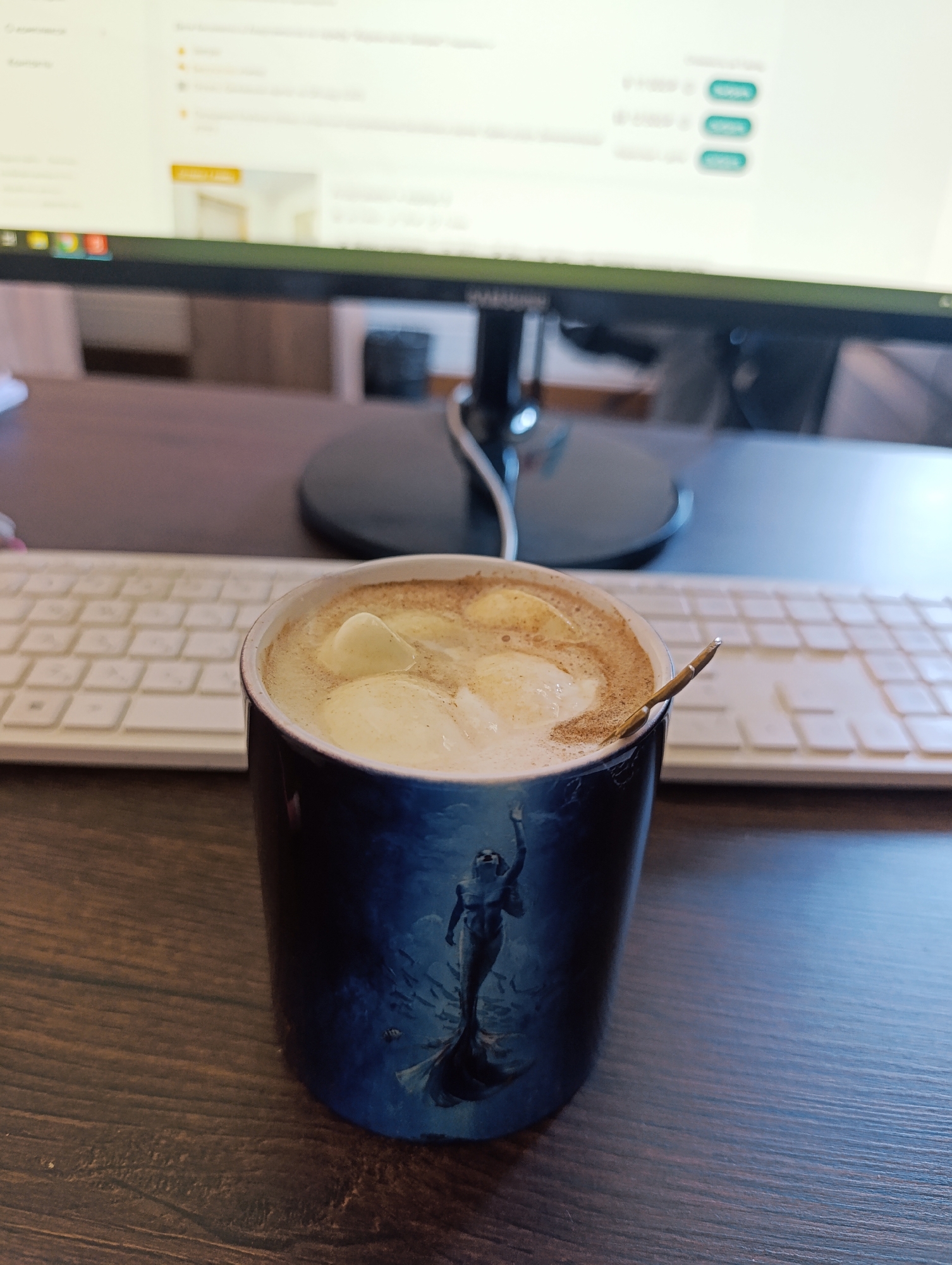 A close-up, slightly high-angle shot shows a dark blue mug with a mermaid design on a wooden desk. The mug is filled with a frothy beverage topped with marshmallows and cinnamon. A small spoon rests on the rim of the mug, partially submerged in the drink. In the background, a white keyboard is visible, followed by a computer monitor displaying a webpage with several green buttons. The lighting suggests an indoor setting, possibly an office or home study.