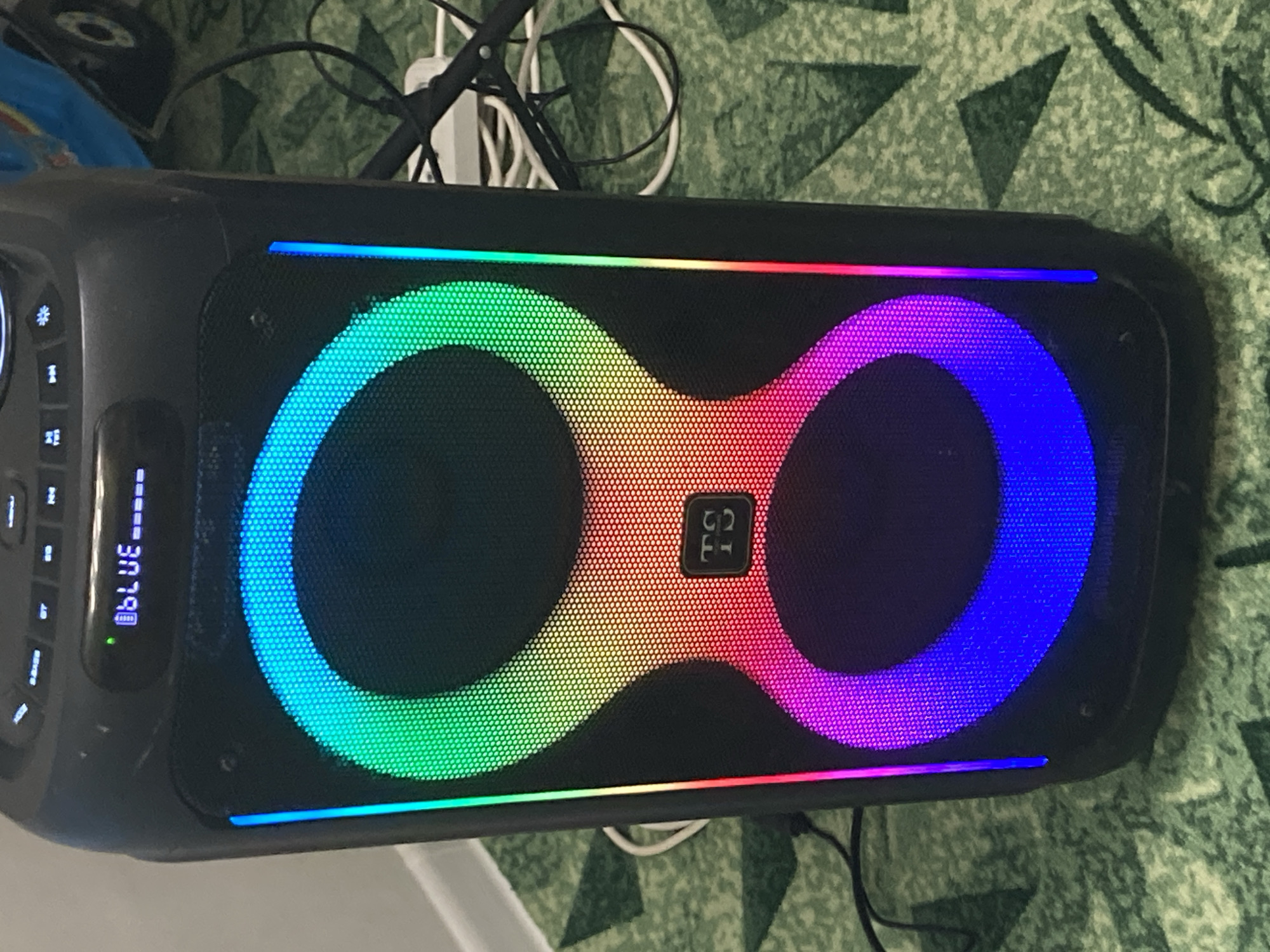 An outdoor portable speaker is shown in a close-up, vertical shot. The speaker is black and rectangular with a large speaker grill in the center. The grill has a colorful LED light display that cycles through shades of green, yellow, orange, red, pink, and blue in a figure-eight pattern. A small, square logo with the letters "TS" is visible on the speaker grill, near the center. The top of the speaker has several buttons and a small digital display that reads "BLUE-----". The speaker is resting on a green carpet with a repeating pattern of lighter green shapes. Wires and cables are visible behind the speaker, connected to what appears to be a power strip. The lighting suggests an indoor environment, possibly a room in a dwelling. There is no visible indication of the city of Chaikovsky, Russia, in the image.