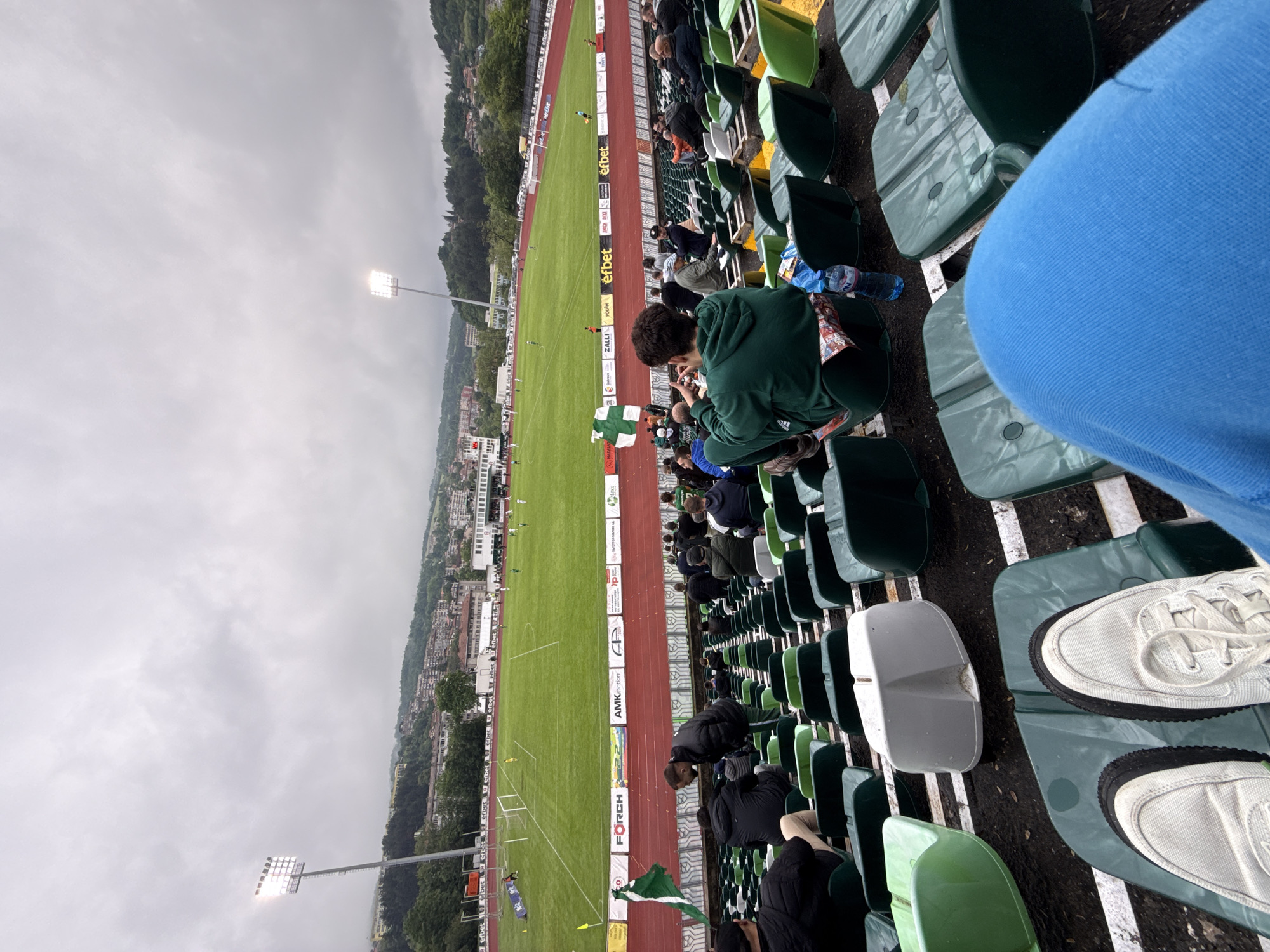 The image captures a view from stadium seating during a soccer game in Gabrovo, Bulgaria. The view is from an elevated perspective, overlooking the soccer field. The field is green and marked with white lines, with the goalposts visible at one end. A red track runs alongside the field, and advertisements in Bulgarian are displayed along the side of the track. Several people are seated in the stadium seating, observing the game. Some are standing or crouched in their seats. The weather appears overcast and gray. Gabrovo's urban landscape stretches out beyond the stadium. Evidence of water is on the seat between the spectator with white sneakers and a boy in the green hoodie who is squatting in his seat taking a photo.