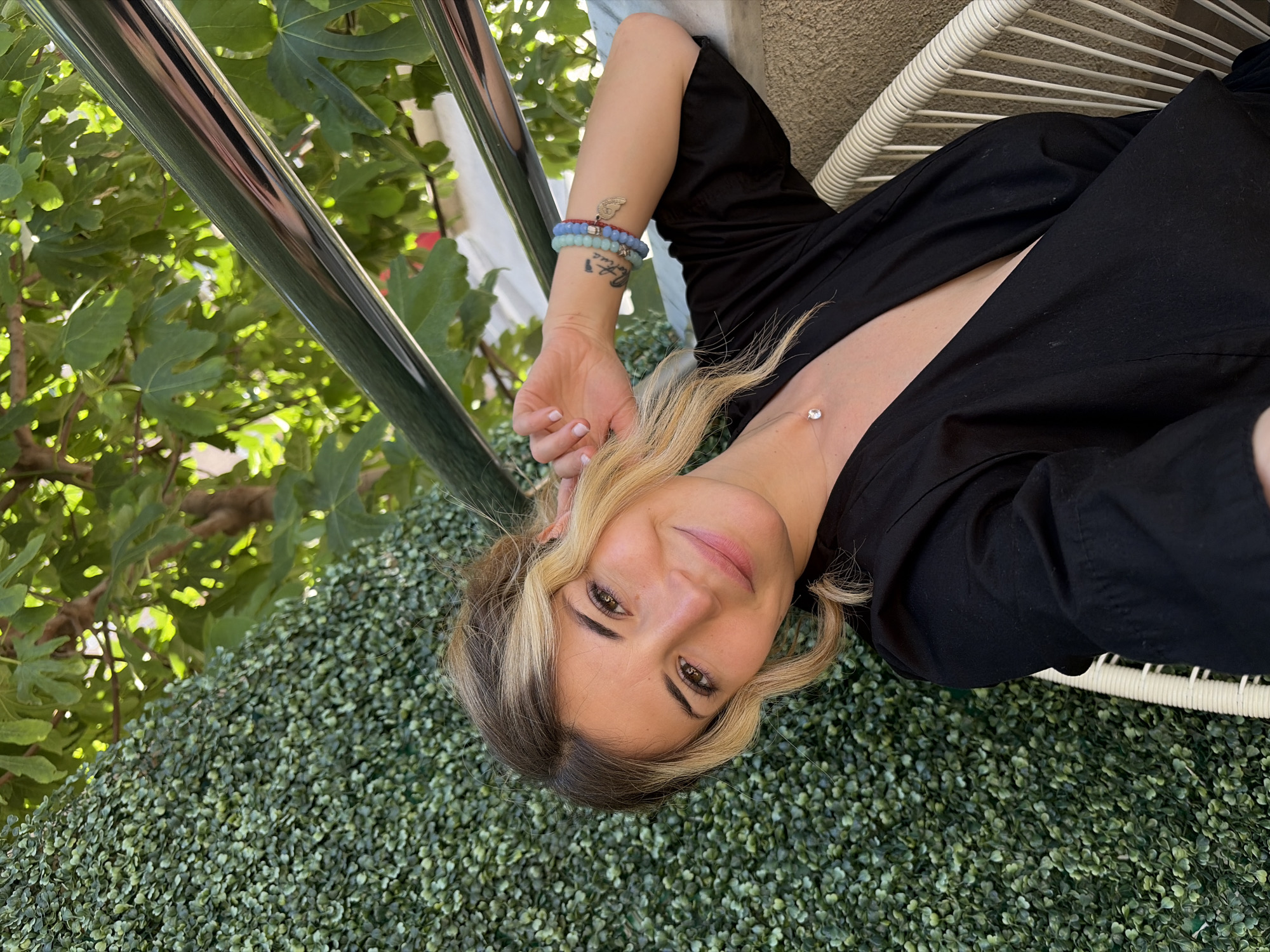 A woman lies at an angle in a white wicker chair on a green backdrop, possibly artificial vegetation. She is wearing a black shirt and has blonde hair with dark roots. She has a diamond or clear stone near her clavicle and bracelets on her left wrist. A metal railing and leaves are visible in the upper left of the frame. The location appears to be outdoors, possibly on a balcony or patio. Though there is no definitive evidence, the location could be Budva, Montenegro.