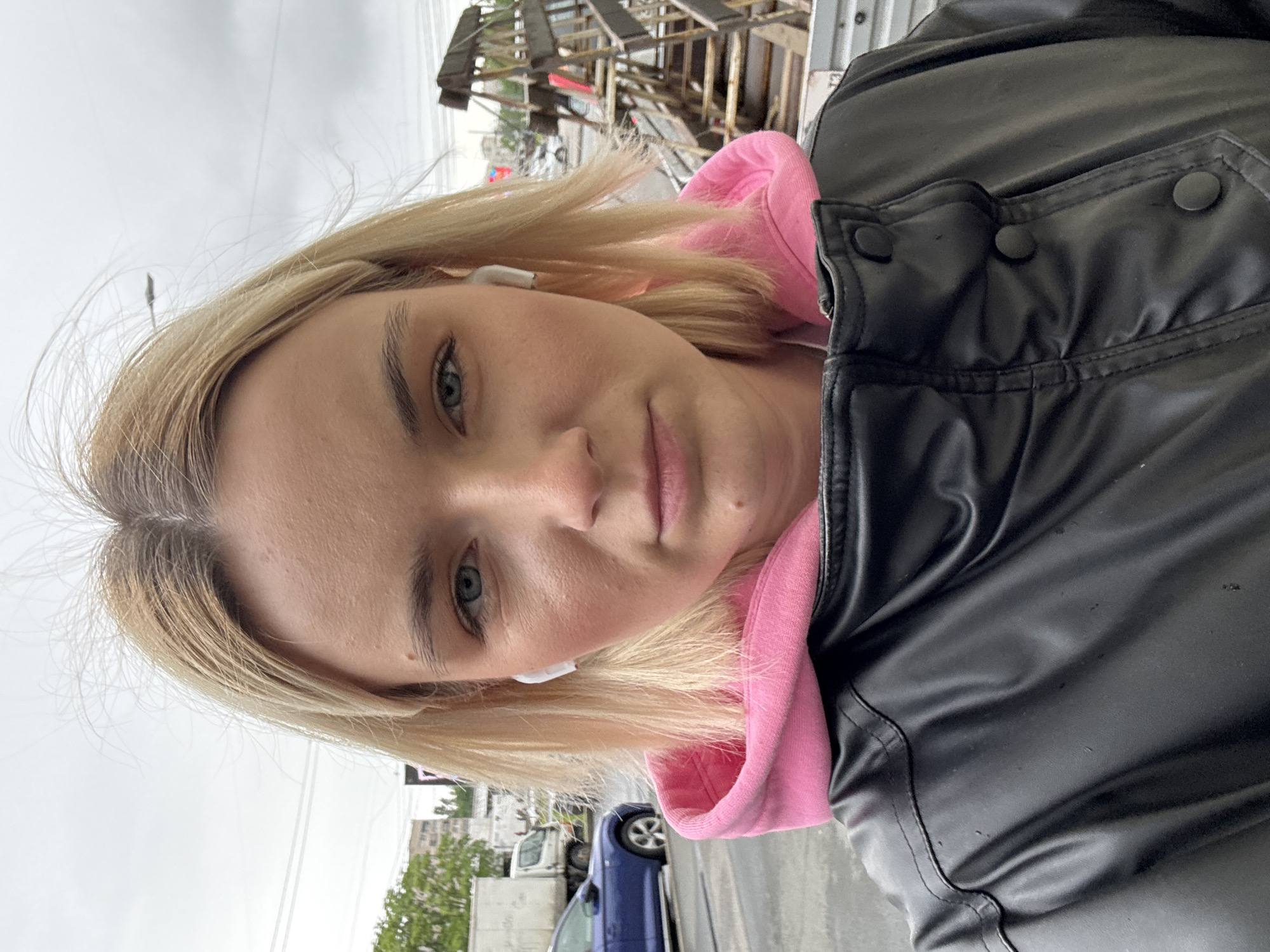 A woman stands outside in what appears to be Saint Petersburg, Russia. She is wearing a black jacket over a pink hooded sweatshirt and has blonde hair. She has wireless earbuds in her ears. In the background, there are buildings, vehicles, and some construction or demolition materials. The sky is overcast.