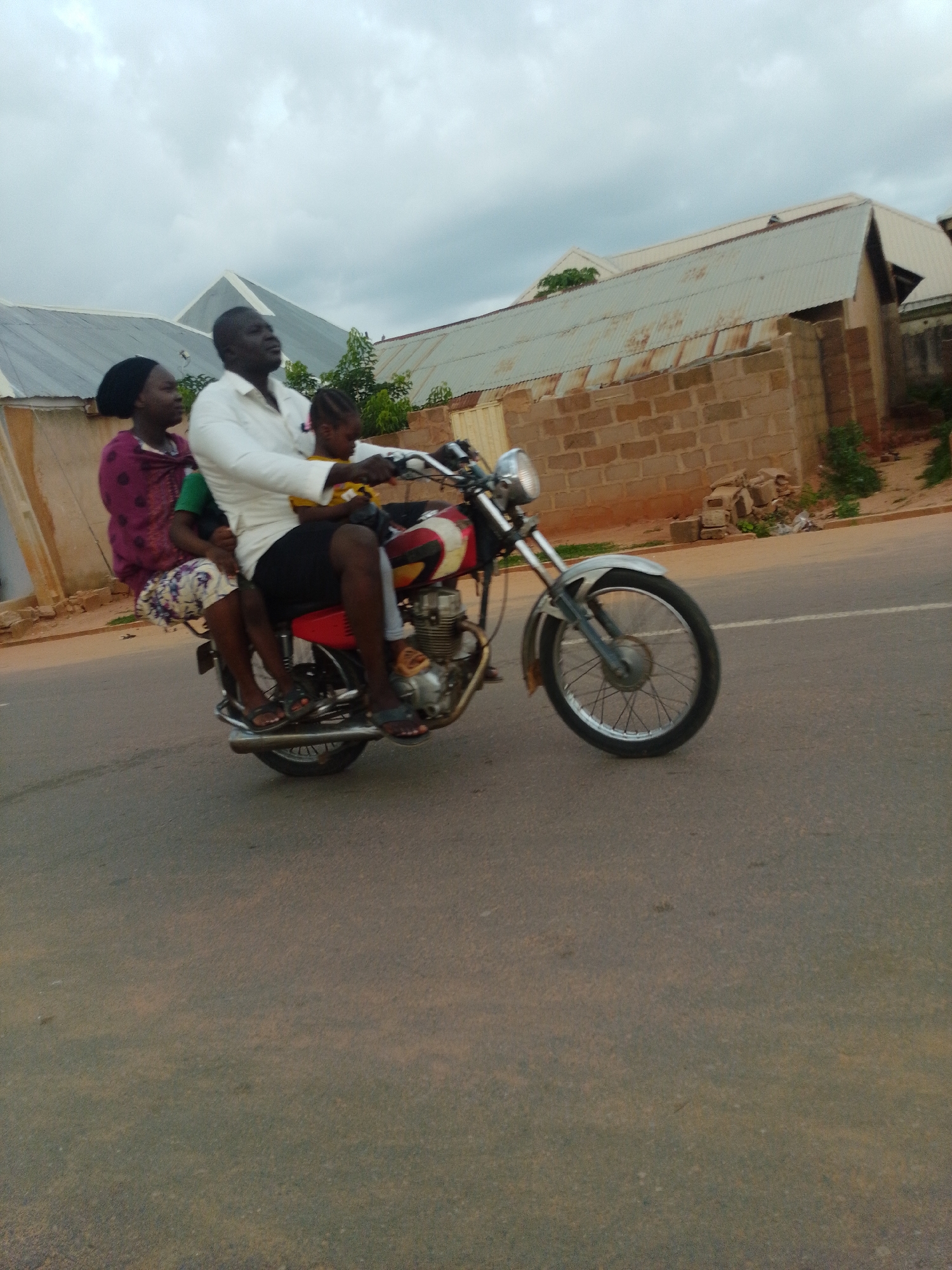 This is a medium shot, eye-level, outdoor photograph taken on a cloudy day in Tempure, Nigeria. The primary subject is a man riding a motorcycle with two passengers. The man is wearing a white long-sleeved shirt and black shorts. He is looking to his left. Seated behind him is a woman wearing a purple garment and a headscarf. In front of the man, between him and the woman, is a young child wearing a yellow shirt. The child is facing forward and looking down. The motorcycle is red and black with silver accents and appears to be moving. The background consists of a road with a white line, some vegetation, and buildings with corrugated metal roofs. One building in the background is made of brick. The overall atmosphere is mundane and typical of daily life in the city.