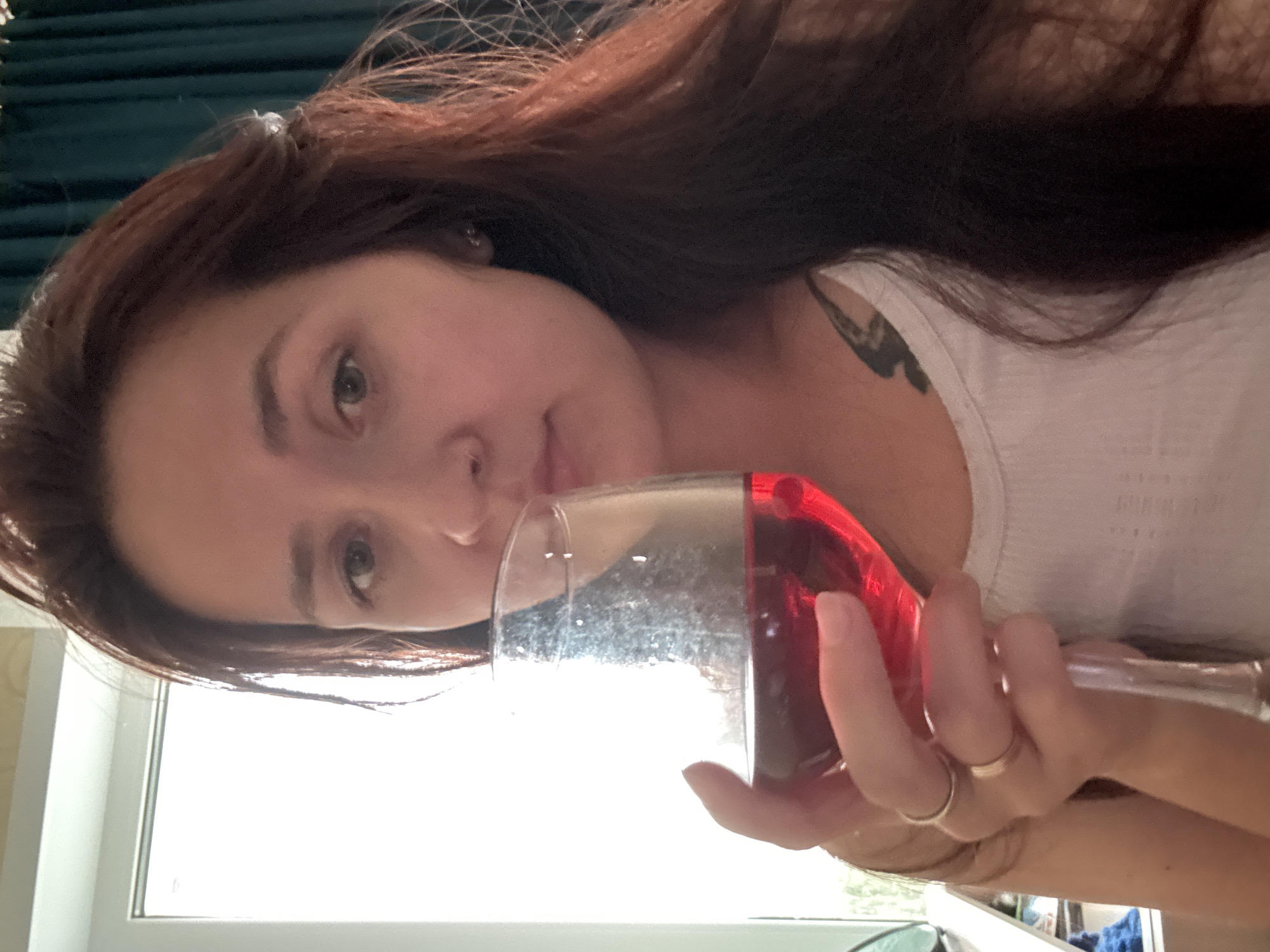 A young woman with long, dark auburn hair is captured in a close-up, selfie-style shot, holding a large wine glass filled with a red beverage. She is wearing a white tank top and two rings on her left hand, which is holding the stem of the glass. A tattoo is visible on her upper chest. Her gaze is directed towards the camera, and her lips are slightly parted. The background is softly blurred, with a bright window on the left and dark teal curtains on the right. There is no specific indication of her location within Naberezhnye Chelny, Russia.
