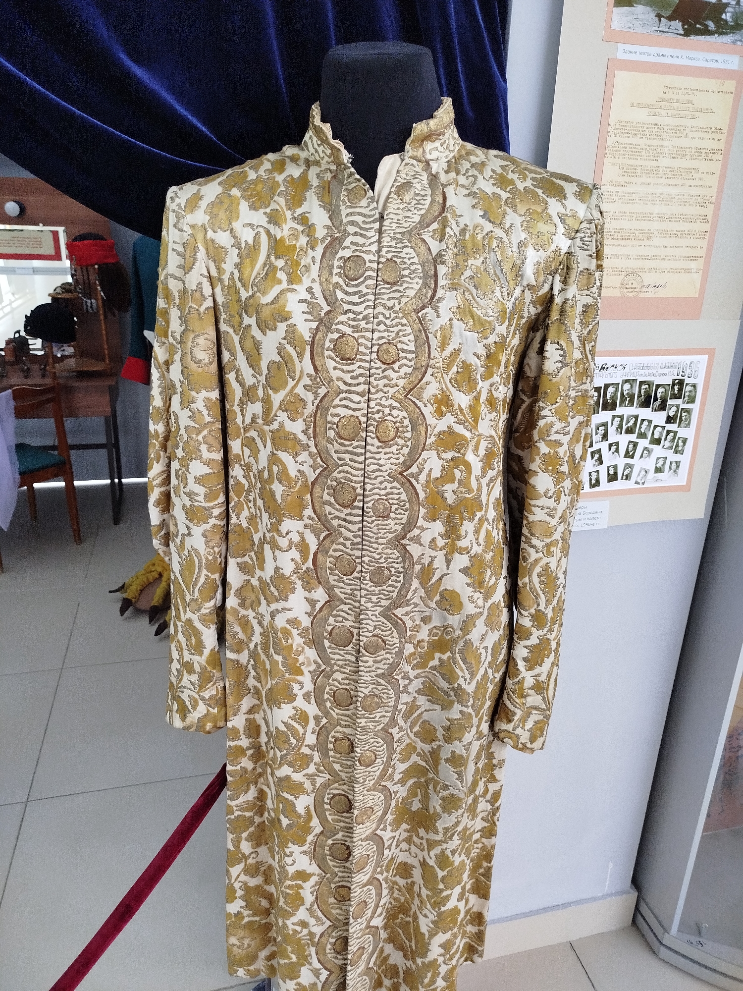 The image shows a display in what appears to be a museum or exhibition. The central focus is a male mannequin dressed in a long, ornate garment. The garment is a richly patterned robe, likely made of silk or brocade, with a white base fabric and a swirling gold floral motif. The robe features a high, standing collar and a front closure with decorative circular buttons or embellishments running down the center.
To the left of the mannequin, a dark blue velvet curtain drapes from above. In the background, a wooden table with chairs is visible, suggesting a room setting. On the table, there are a few items, including what looks like a red hat. On the wall behind the mannequin, two framed exhibits are displayed. The top exhibit is a document with text and a small image, while the bottom exhibit is a collage of black and white photographs of people. A red velvet rope barrier is positioned in front of the display. The floor is tiled in a light color. The overall impression is of a curated exhibit of historical clothing and artifacts.