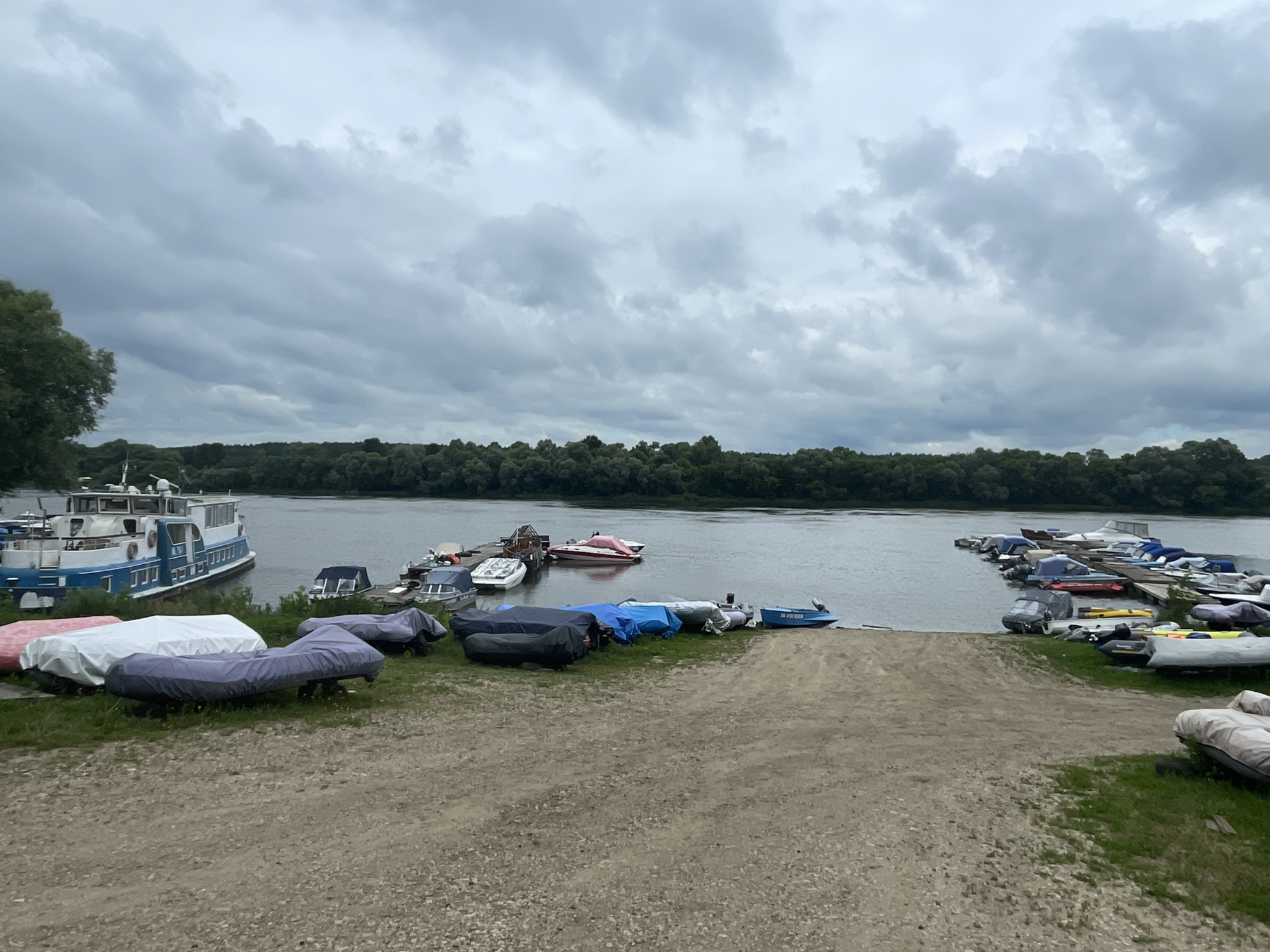 The image depicts a cloudy day at a boat dock on the Oka River in Pushchino, Russia. Several boats of various sizes are moored or stored along the shore and on a wooden pier. A large blue and white passenger boat is docked on the left side of the image. Numerous smaller boats, some covered with tarps and others visible, are arranged on the grass and near the water's edge. A sandy ramp leads down to the river. The background features a dense line of trees along the opposite bank of the river, under a heavily overcast sky. There are no people visible in the scene.