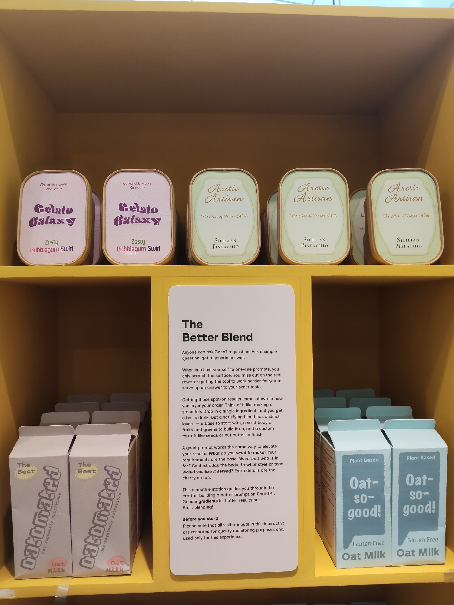 The image depicts a display shelf at an interactive exhibit in Singapore. On the top shelf, there are four cylindrical containers of gelato, two labeled "Gelato Galaxy" with "Zesty Bubblegum Swirl" flavor and two labeled "Arctic Artisan" with "Sicilian Pistachio" flavor. The bottom shelf contains two rows of carton-like products. The front row shows two "Datonated Oat Milk" cartons with "Oat-rageously nutritious" printed on them. The back row shows "Oat-so-good!" cartons labeled "Plant Based" and "Gluten Free Oat Milk". A yellow sign to the right of the bottom shelf, titled "The Better Blend," provides instructions for using a chatbot to create smoothie blends. The overall environment appears to be a clean and well-lit indoor exhibition space.