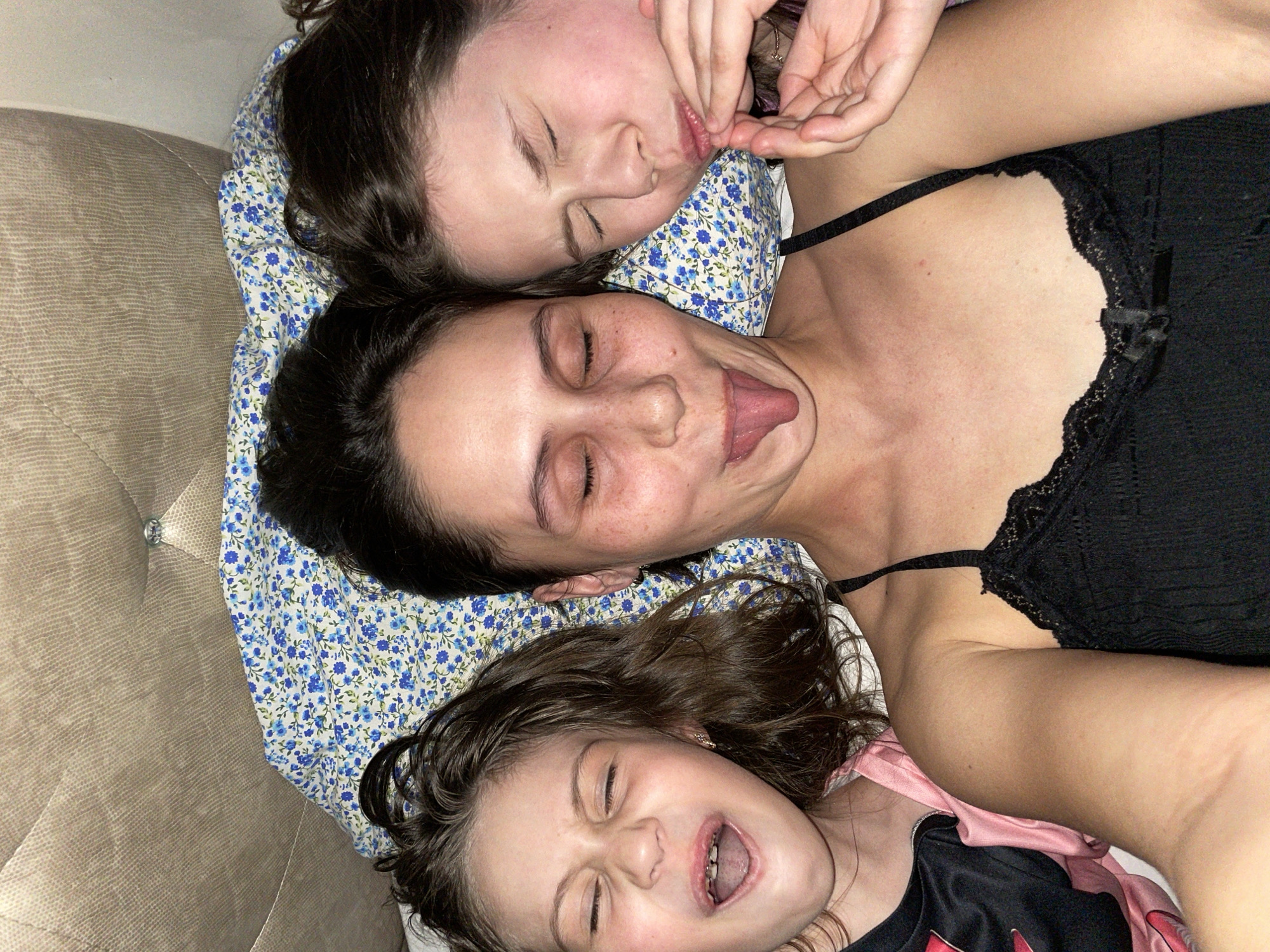 Three females, likely family members, are captured in a playful moment, lying close together on what appears to be a bed or sofa. They are positioned with their heads aligned, facing upwards towards the camera.
The topmost individual, with dark hair, has her eyes closed and her lips pursed, with one hand seemingly touching her face. Below her, a second woman, also with dark hair and freckles, has her eyes closed and her tongue playfully sticking out. She is wearing a black lace camisole. The third and youngest individual, positioned at the bottom of the frame, has dark curly hair, her eyes squeezed shut, and her mouth wide open in a loud laugh or shout. She is wearing a pink and black top.
Their heads rest on a pillow with a blue floral pattern. Behind them, a beige, textured upholstered surface, possibly a headboard or couch back, is visible. The lighting is soft and warm, suggesting an indoor setting, likely in the evening or night. The overall mood of the image is one of joy, playfulness, and familial intimacy. There is no visible text in the image. The location context provided is Izhevsk, Russia.