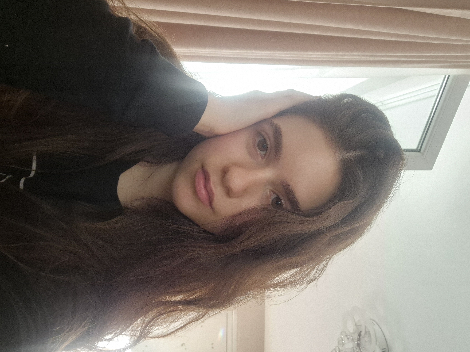 A young woman is facing the camera in a portrait. She has long brown hair, light skin, and neutral makeup. Her left hand is resting on her head. She's wearing a black top. Behind her are a window and blinds. There's no information to suggest this image was taken in Pavlikeni, Bulgaria.