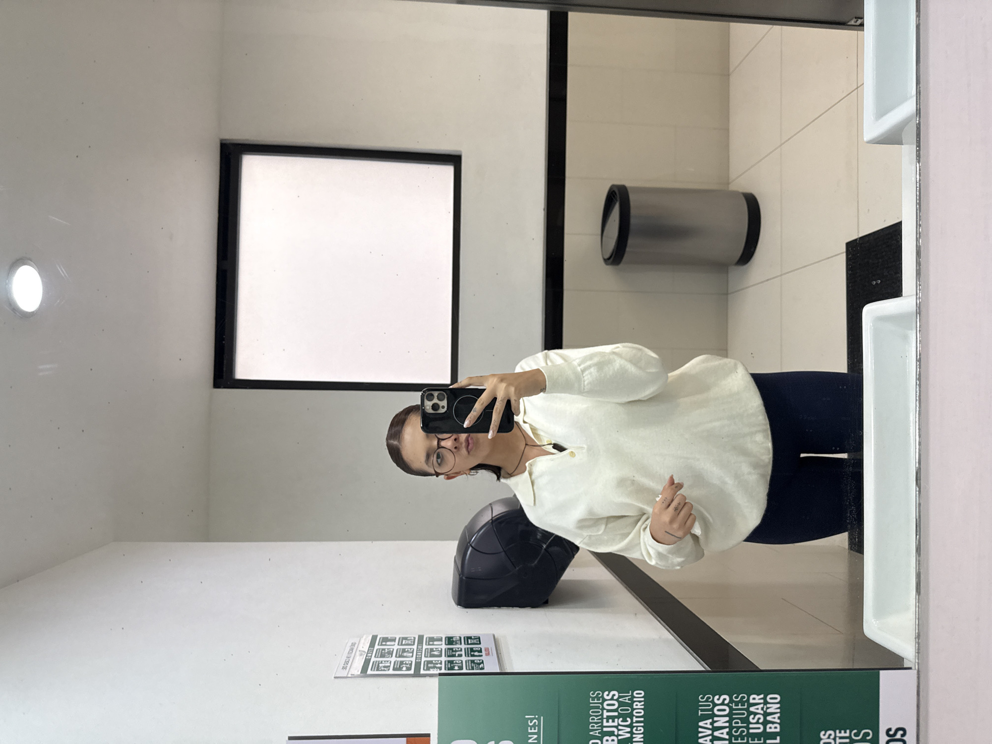 A woman is taking a mirror selfie in what appears to be a public restroom. She is holding a smartphone, wearing glasses, a light-colored long-sleeved shirt, and dark pants. The restroom has white walls, a black-framed window, a hand dryer, and a black trash receptacle. A sign in Spanish regarding proper restroom use is partially visible in the foreground. The restroom appears to be in San Andrés Cholula, Mexico, based on the Spanish signage.