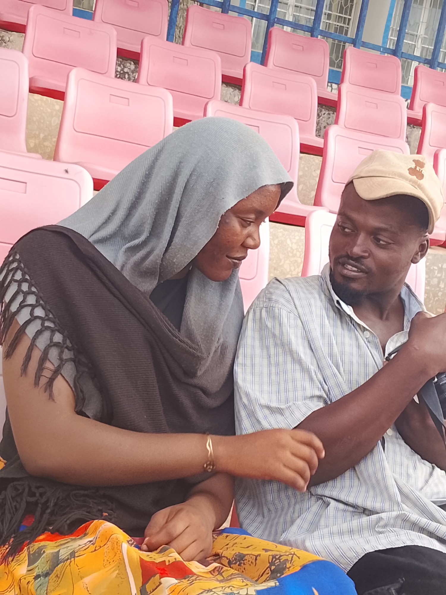This media file depicts two individuals, a woman and a man, seated within what appears to be a stadium or public venue in Zawan, Nigeria. The background features several rows of empty, vibrant pink plastic seats, with blue structural elements visible further back.
The woman, on the left, is wearing a light gray head covering (possibly a hijab or khimar) and a dark gray or black shawl with fringed edges draped over her shoulders. A brightly patterned fabric, featuring yellow, orange, blue, and red, is visible on her lap. She wears a simple gold-colored bracelet on her right wrist. Her gaze is directed towards the man with a soft, engaged expression.
The man, seated on the right, wears a light-colored, vertically striped short-sleeved button-up shirt and a beige baseball cap with a dark, unidentifiable emblem on the front. He appears to be holding a camera or similar device in his right hand. He is looking at the woman with an expressive, engaged facial expression, as if in mid-conversation.
The scene suggests a relaxed, informal interaction between the two individuals. The even lighting indicates it is daytime, possibly under an overcast sky or within a shaded area of the venue. The empty seats imply they are either early arrivals, attending a less crowded event, or perhaps at a practice or casual gathering. There is no visible text apart from the indistinct emblem on the man's cap.
