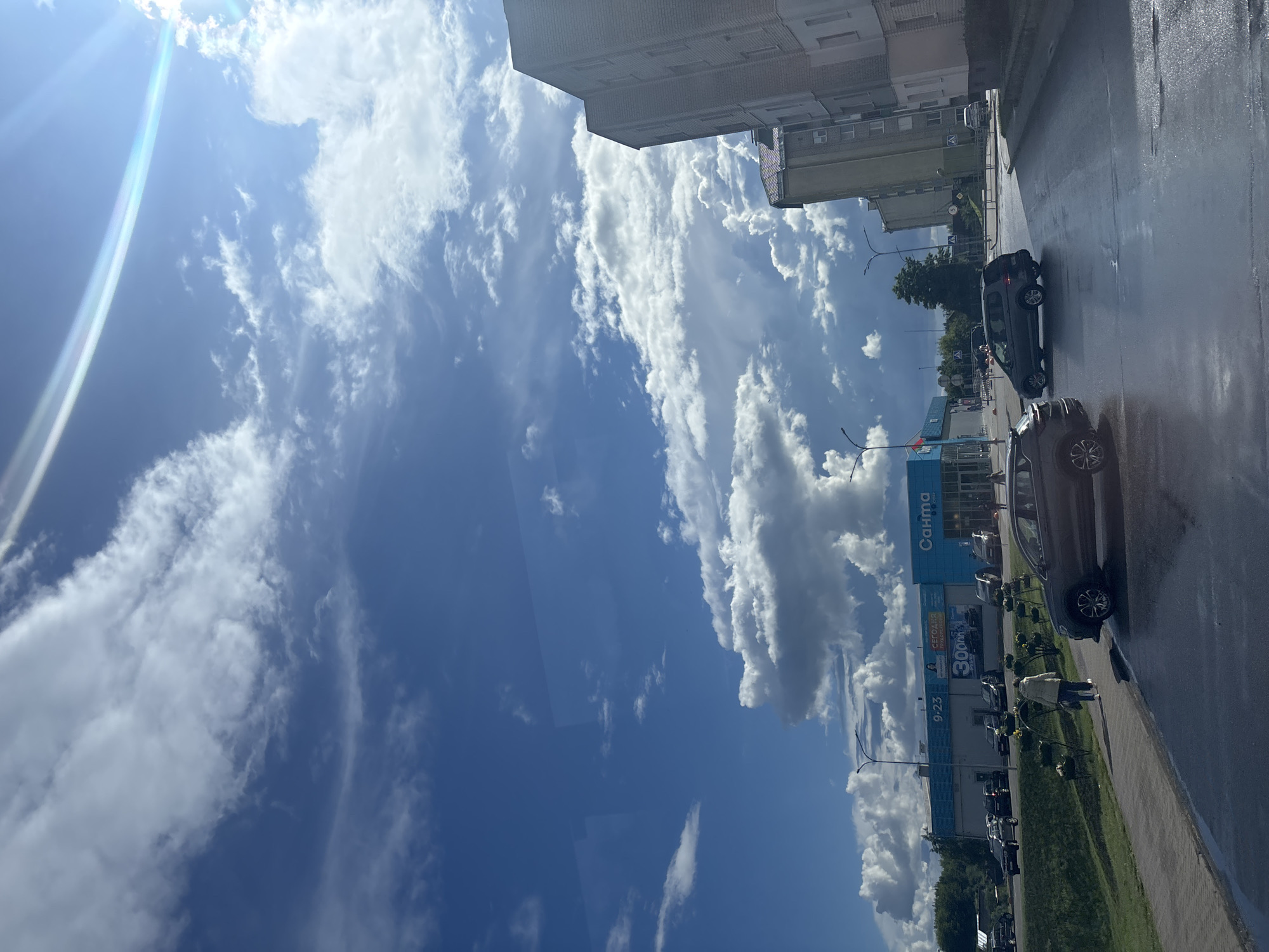 The image captures a street scene in Kapyĺ, Belarus, under a dramatic sky filled with both bright sunlight and large, dark cumulus clouds. In the foreground, a wet asphalt road reflects the sky, with two cars visible on it: a dark gray SUV on the left and a gray minivan further ahead on the right. A sidewalk runs along the left side of the road, where a woman in a light-colored jacket and dark pants is walking away from the camera, carrying a bag.
To the left, a blue and white supermarket building with a sign reading "Санта" (Santa) is prominent. The store appears to be open, with the hours "9-23" displayed. Several cars are parked in front of the supermarket. Beyond the store, there are green trees and a glimpse of other buildings.
To the right, a tall, multi-story brick apartment building dominates the scene. The weather appears to be transitioning, with the sun creating lens flares in the upper right corner of the image, suggesting a break in the clouds or a shift in weather patterns. The overall atmosphere is one of a typical urban environment on a day with dynamic weather conditions.