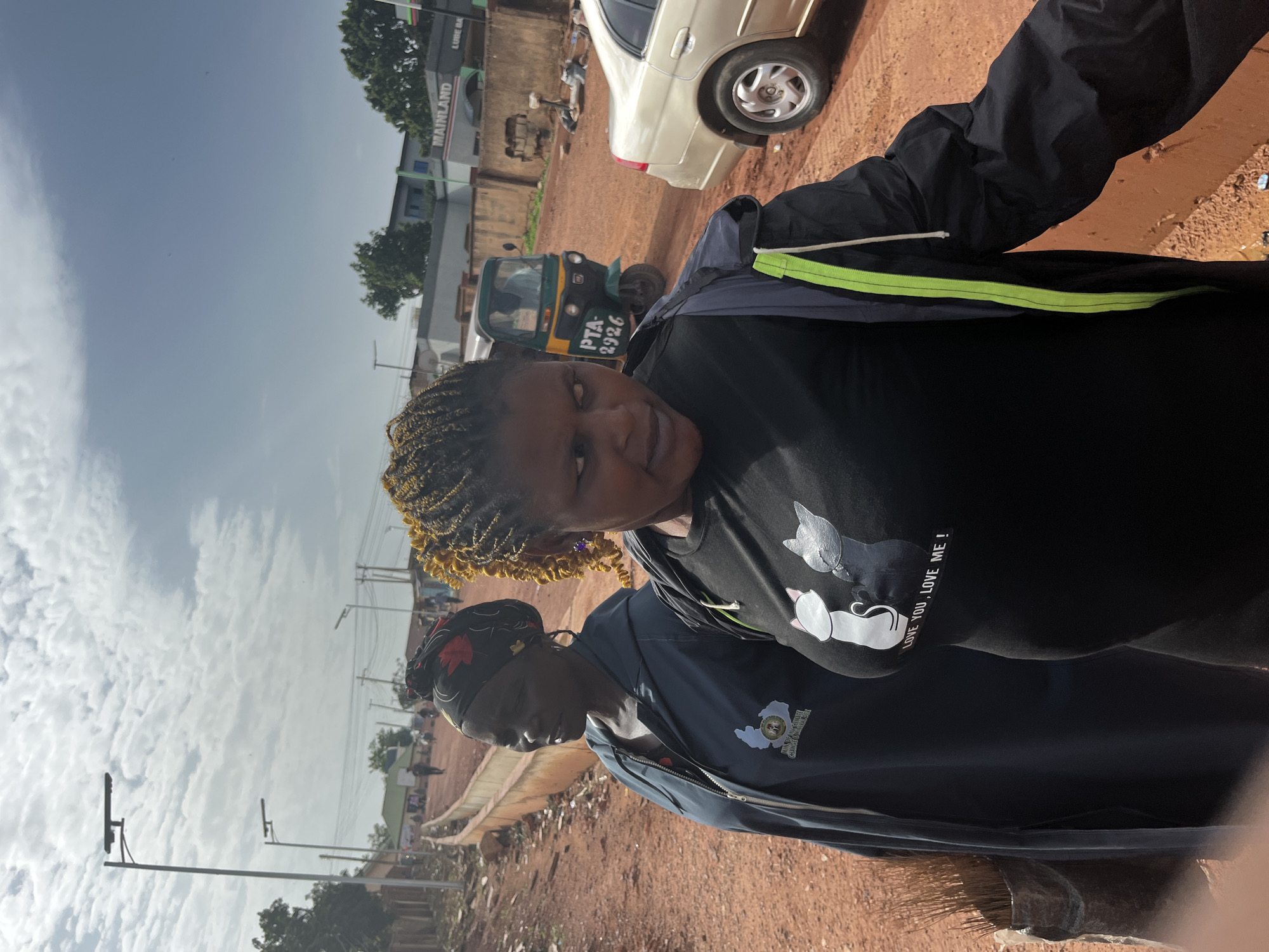 This image depicts an outdoor scene in Gura Top, Nigeria, under a partly cloudy sky. Two women are the primary subjects, positioned in the foreground. The woman on the left wears a dark headscarf with a red and black floral pattern and a dark blue hooded jacket with a logo on the left chest. She is looking to her left. The woman on the right has braided blonde hair and is wearing a black t-shirt with a graphic of two cats and text that reads "LOVE YOU, LOVE ME !". She is looking to her right. She also wears a black jacket with green trim.
The environment is a dirt road with reddish-brown soil. On the left side of the road, there is a raised earthen embankment lined with concrete. In the background, several streetlights are visible, suggesting it is a public road. Buildings line the background, including one with "MAINLAND" signage and another that appears to be a gas station or similar commercial establishment. A three-wheeled auto-rickshaw (also known as a Keke Napep) is visible on the road, along with a light-colored sedan parked on the right. Several other people are visible in the mid-ground, walking along the road. The overall impression is a typical urban or semi-urban street scene in Nigeria.