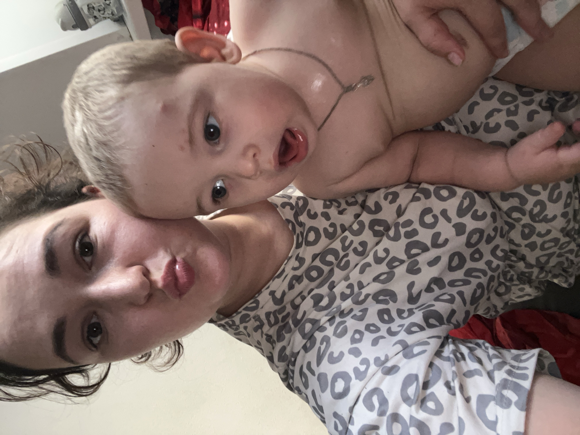 A woman and a baby are posing for a selfie indoors, with the woman's face partially obscured as she makes a kissing gesture. The baby, facing to the right, has an open mouth and wide eyes. Both are dressed casually, with the woman wearing a leopard-print top. The background appears to be a domestic setting. The location of this image is unknown and cannot be determined to be in Chelyabinsk, Russia.