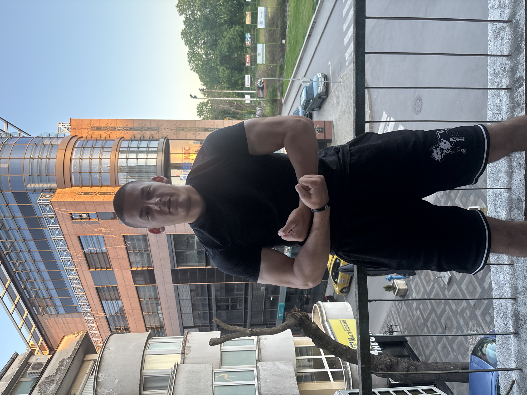 A man stands on a balcony, flexing his bicep muscles. He is wearing a black t-shirt and black shorts with a white trim and an image of two dogs or wolves on the side. He has short, buzzed hair and a wrist watch. The balcony has a metal railing, offering a view of the street below.
The scene includes a building with a glass facade and a bronze-colored exterior. Visible from the balcony are cars, trees, and other buildings. The signage “ЕКТРАДРАВЕ” is visible on a structure below, written in Cyrillic, a part of the local signage. Based on the architecture, color palette of buildings, Cyrillic text and environment, the location is likely Plovdiv, Bulgaria.