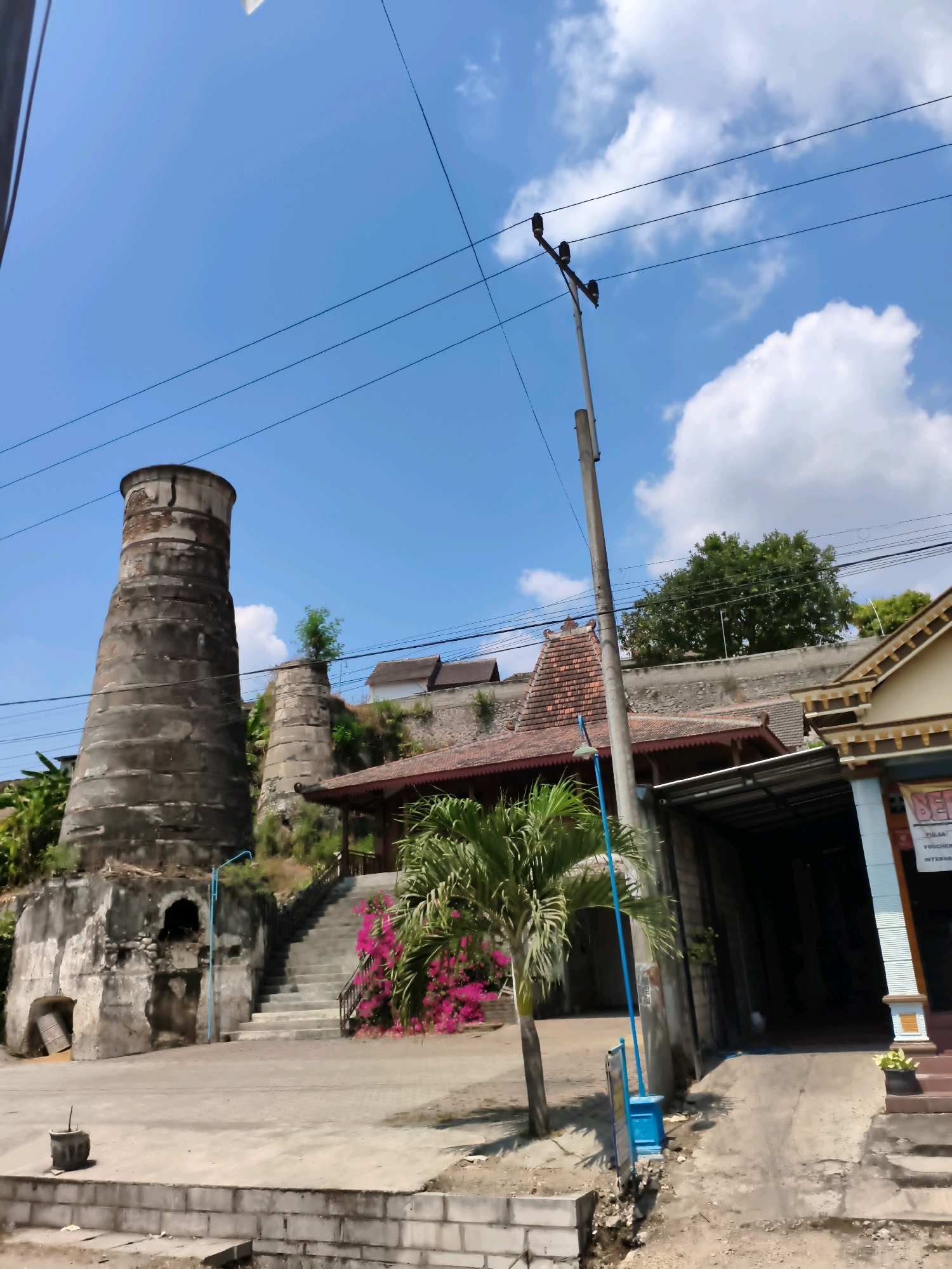 The image depicts an outdoor scene in Sampung, Indonesia, featuring an old, cylindrical, stone structure reminiscent of a kiln or tower on the left. Adjacent to it are a set of stairs leading up to a building with a traditional tiled roof. A palm tree and vibrant pink bougainvillea flowers are visible near the stairs. A utility pole with electrical wires stretches across the frame, and a building with a more modern facade stands on the right side of the image. The sky is blue with scattered white clouds. No people are present in the scene. The overall environment suggests a historical or cultural site within a town or village setting.