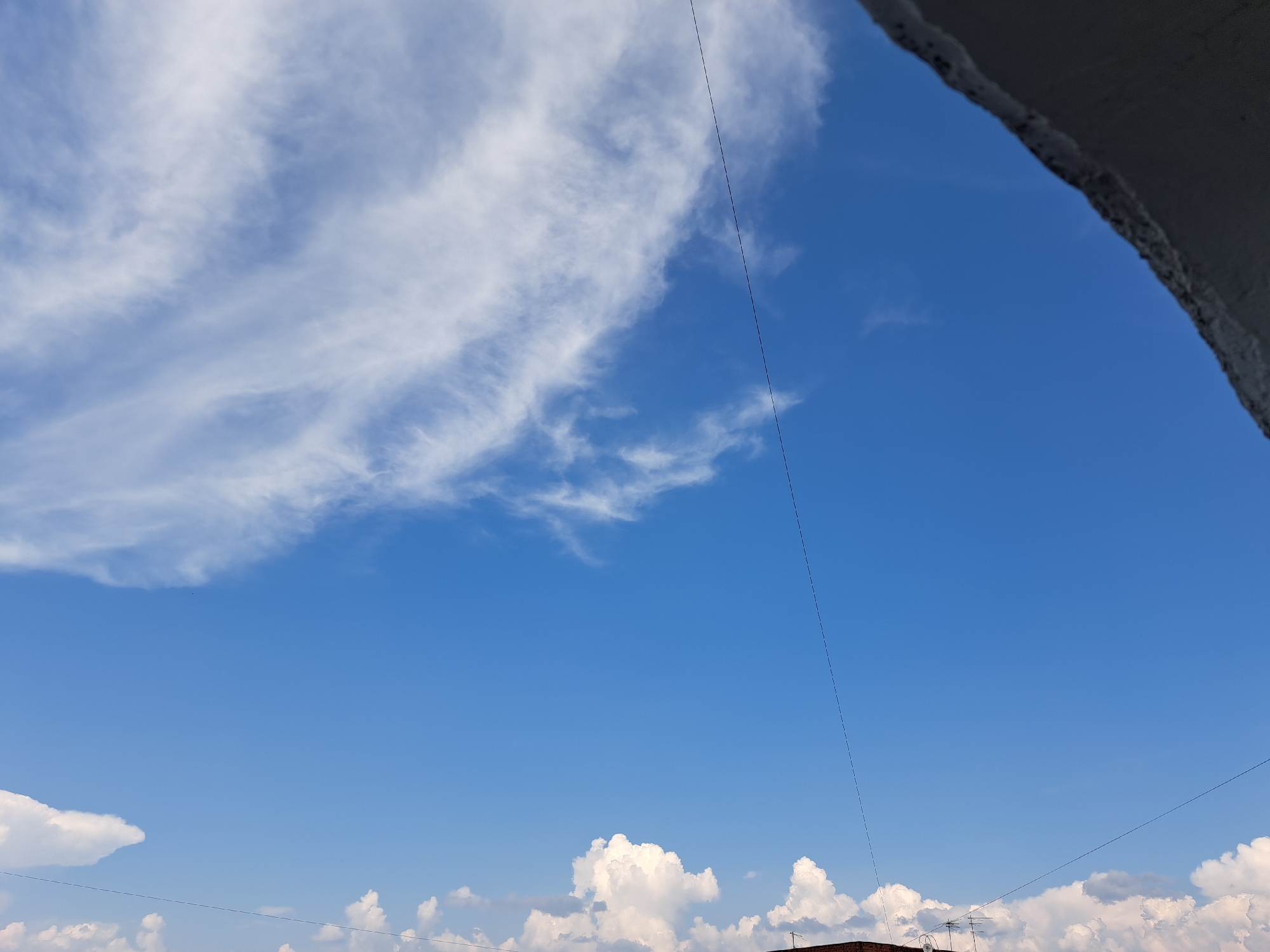 A wide-angle, low-angle shot captures a bright blue sky with scattered white clouds. In the upper right corner, a portion of a dark, rough-textured structure, possibly a rooftop or overhang, juts into the frame. A thin black wire stretches diagonally across the sky from the upper right towards the lower center. In the lower portion of the image, the tops of buildings are visible, suggesting an urban environment. The clouds are wispy and stretched in the upper left, with more dense, cumulus clouds lower down. The overall impression is that of a clear day, likely in a city like Irkutsk, Russia, given the context of your request.