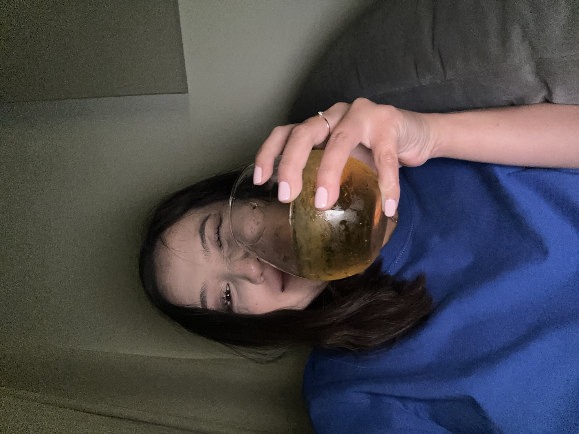 A young woman with dark hair, wearing a blue t-shirt, is holding a wine glass filled with a light-colored liquid and ice. She is looking towards the camera and winking. Her left hand is on the glass, and she has light pink nail polish. A thin ring with a small blue stone is visible on her ring finger. The background is dimly lit and appears to be indoors, with a draped fabric on the left and a muted-colored surface on the right. The scene is set in a dimly lit room, possibly a home, with a casual atmosphere. There is no discernible location in North Kuta, Indonesia.