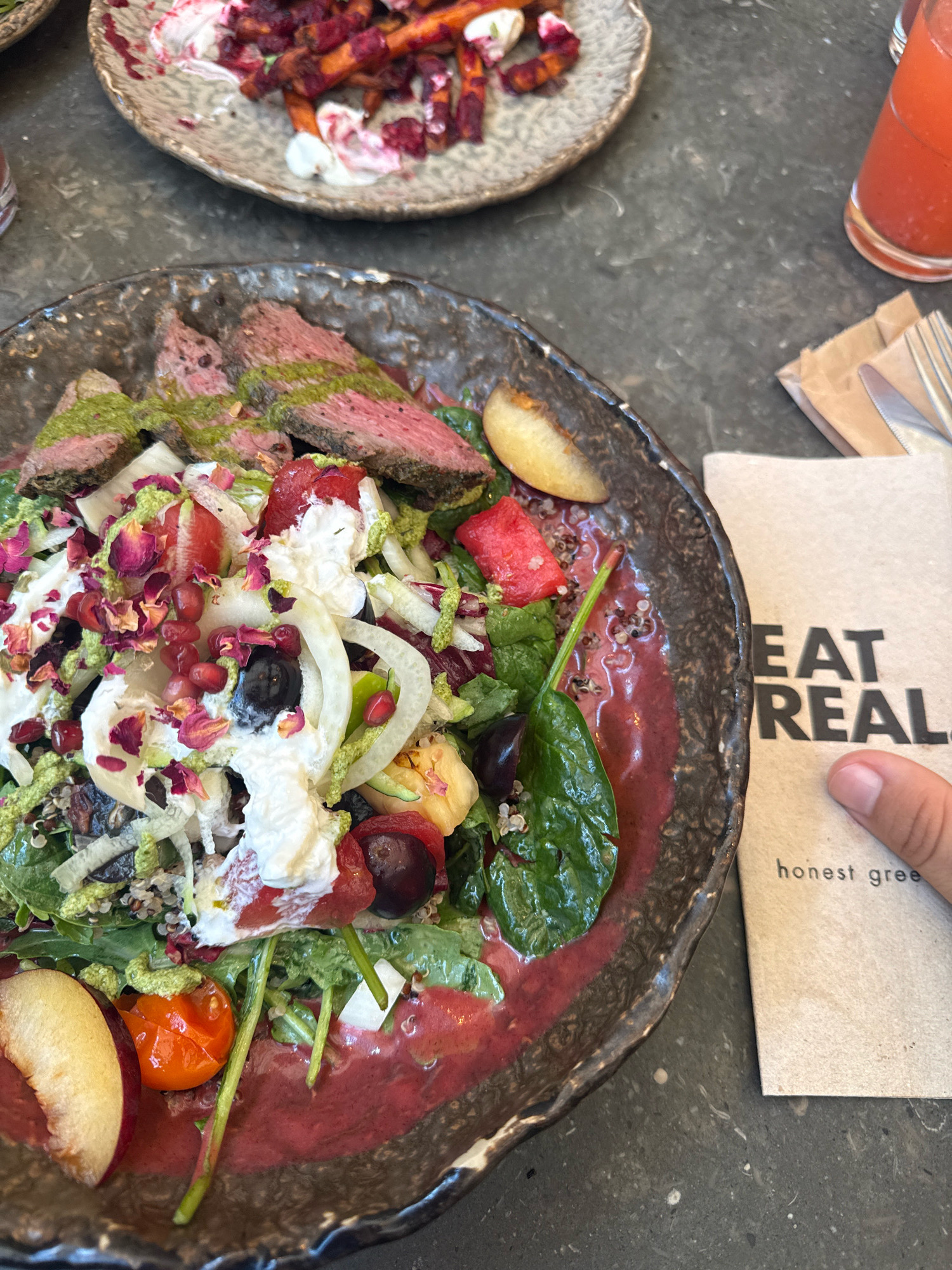 A close-up overhead shot shows a steak and salad dish on a dark, textured surface. The salad is in a rustic, dark-rimmed bowl and features sliced steak, mixed greens, quinoa, white cheese, pomegranate seeds, olives, sliced peaches, and cherry tomatoes, all drizzled with a green sauce and a vibrant red dressing. To the right of the salad, a folded brown napkin with black lettering reads "EAT REAL" and "honest gree". A hand is visible on the right side of the frame, pointing at the napkin. In the upper background, another plate holds roasted vegetables, likely carrots and beets, with white sauce. A tall glass of red liquid, possibly juice, is also visible in the upper right. The setting appears to be a restaurant in Porto, Portugal, given the presence of the napkin, suggesting a contemporary dining establishment.