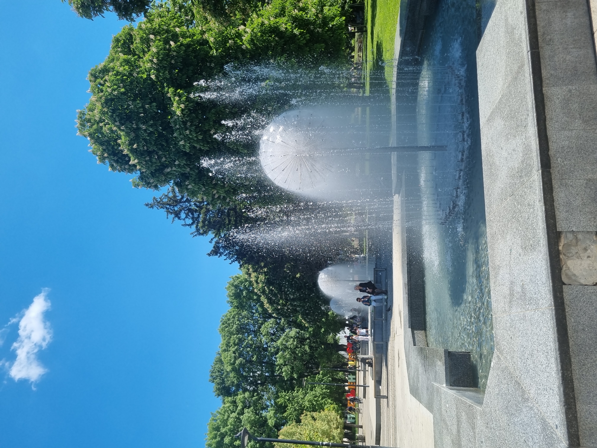 The image depicts a fountain in Pleven, Bulgaria, with a spherical spray pattern creating a dandelion-like effect. Water is visible in a pool, with gray stone surrounding it. Trees line the background, contrasting with a bright blue sky with one small cloud. People are near the fountain, suggesting a public space. A paved area is also visible in the foreground. Benches and lampposts are also present along the paved area.