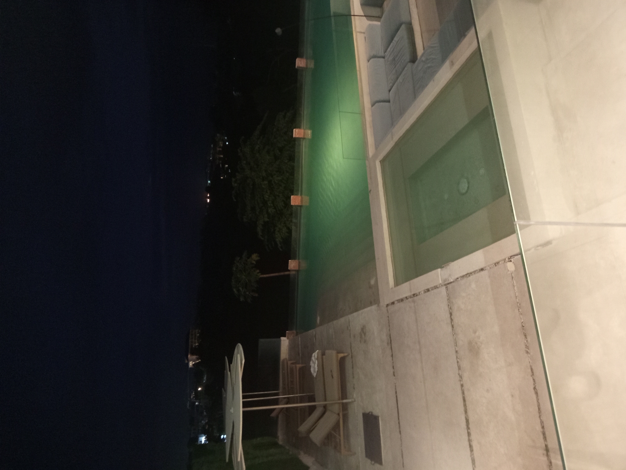 The image depicts an outdoor setting at night, possibly in Kuta Selatan, Indonesia, based on the environment. To the left, a body of water reflects the dark night sky. Moving right, the image shows foliage and a palm tree, suggesting a tropical location. A swimming pool, illuminated with a green light, runs through the center of the image. Adjacent to the pool, there's a seating area with cushioned benches. Further to the right, the structure appears to be a building with a window or glass wall. On the lower part of the image, there are what appear to be clothes drying racks and loungers on the lawn, adding a sense of residential or resort-like activity. The image is oriented sideways.