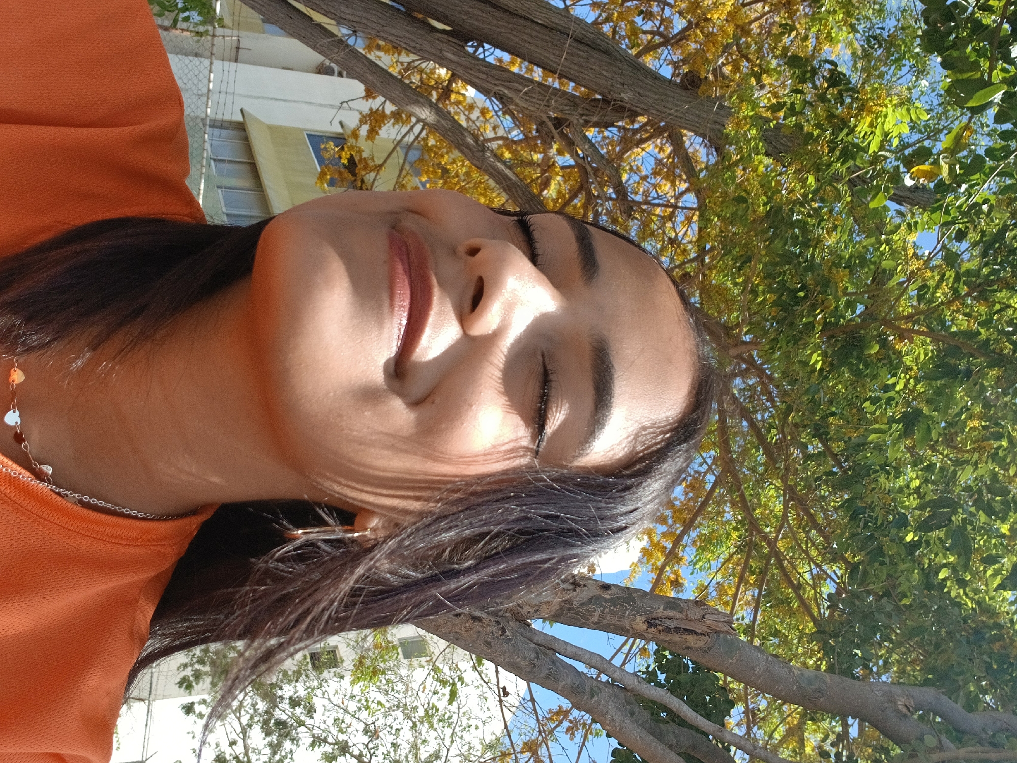 A woman with brown hair, wearing an orange shirt and a silver chain necklace, is shown from a low angle, looking upward with her eyes closed. The background features tree branches with green and yellow leaves against a blue sky. Parts of a building with white walls and windows can also be seen through the branches. The location is possibly Altagracia, Venezuela, although no specific details confirm this.