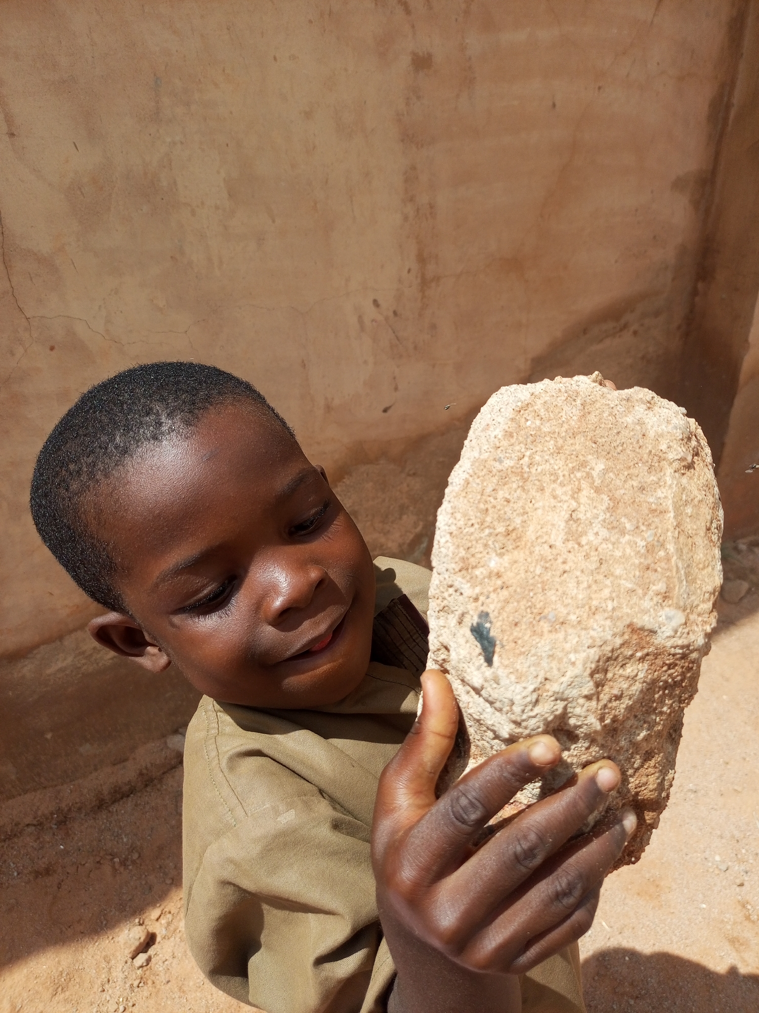 In the city of Gombe, Nigeria, a young Black boy with short, dark hair and brown skin is shown from the chest up, looking down at a large, light-brown rock he is holding in his hands. The boy is wearing a tan shirt. The rock is rough and textured, with some dark patches on its surface. He is smiling slightly and appears to be examining the rock closely. The background is a textured, sandy-colored wall. The lighting suggests an outdoor setting, likely during the day, with shadows indicating sunlight. There are two small, dark insects flying near the rock.