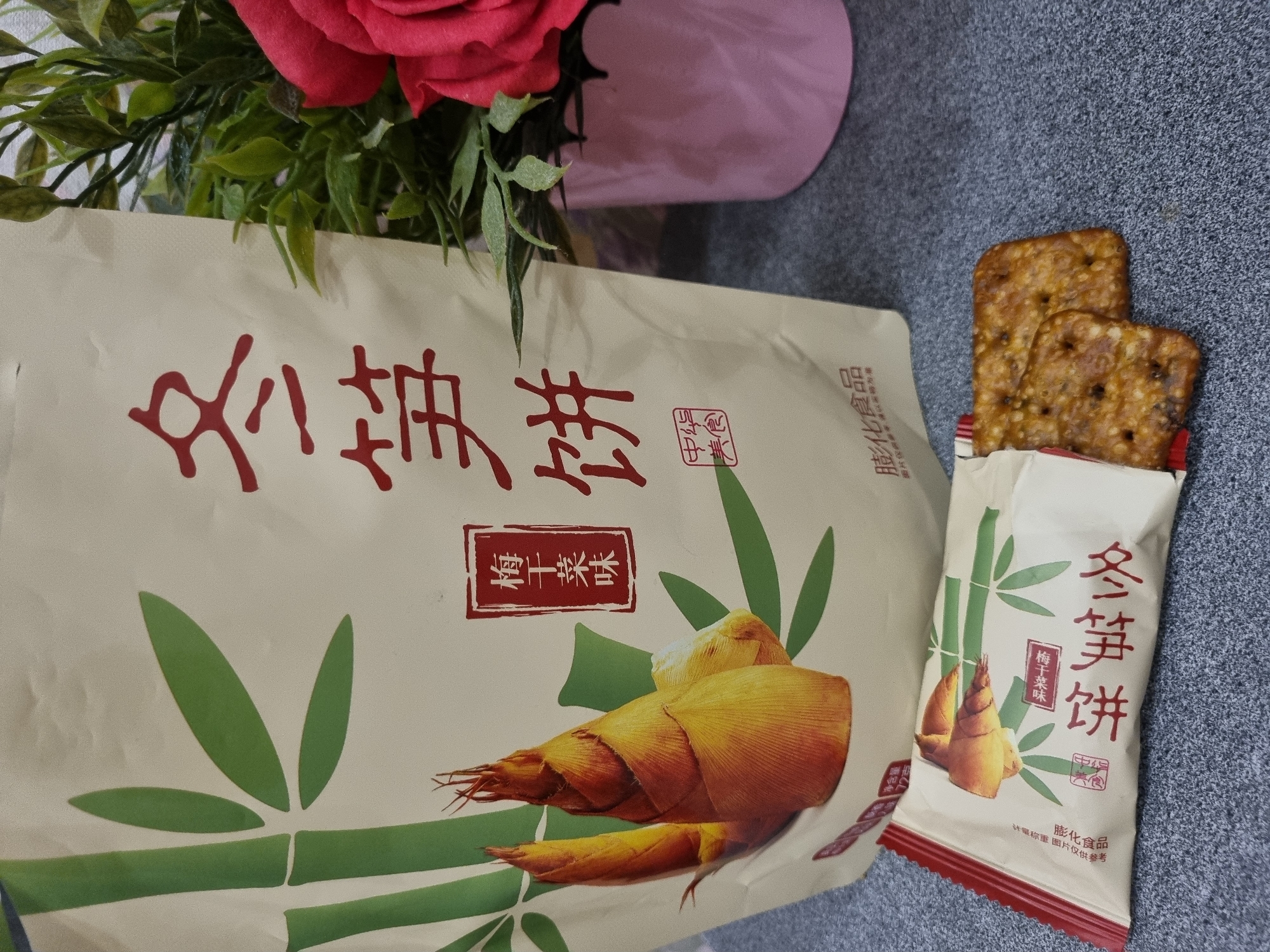 The image shows two packages of "Winter Bamboo Shoots" crackers, one larger package lying on its side and a smaller package standing upright with two crackers protruding from the top. The packages have a cream-colored background with depictions of bamboo and bamboo shoots. The Chinese characters "冬笋饼" are prominently displayed on both packages. The smaller package also has the characters "梅干菜味" (dried plum flavor). A bouquet of red roses is partially visible in the upper left corner of the image. The crackers are dark brown with small holes or indentations. The surface below the crackers and packages appears to be a gray countertop or tabletop.