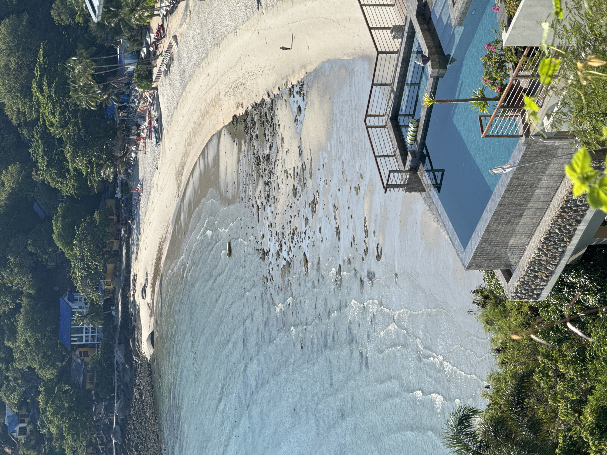 An aerial view captures a serene coastal scene with a crescent-shaped sandy beach meeting the turquoise ocean. A luxurious infinity pool with dark tiled edges is in the foreground, overlooking the beach and sea. The pool area is surrounded by a wooden deck and railings, with a small palm tree and flowering bushes providing decorative elements. Beyond the beach, lush green trees and several houses are visible on a hillside, with one house featuring a distinctive bright blue roof. The beach itself is sparsely populated, with a single person walking along the shoreline. The water exhibits gentle waves and shows rocky formations near the shore, indicating a shallow depth in these areas. The scene is bathed in bright sunlight, creating a peaceful and idyllic tropical atmosphere.