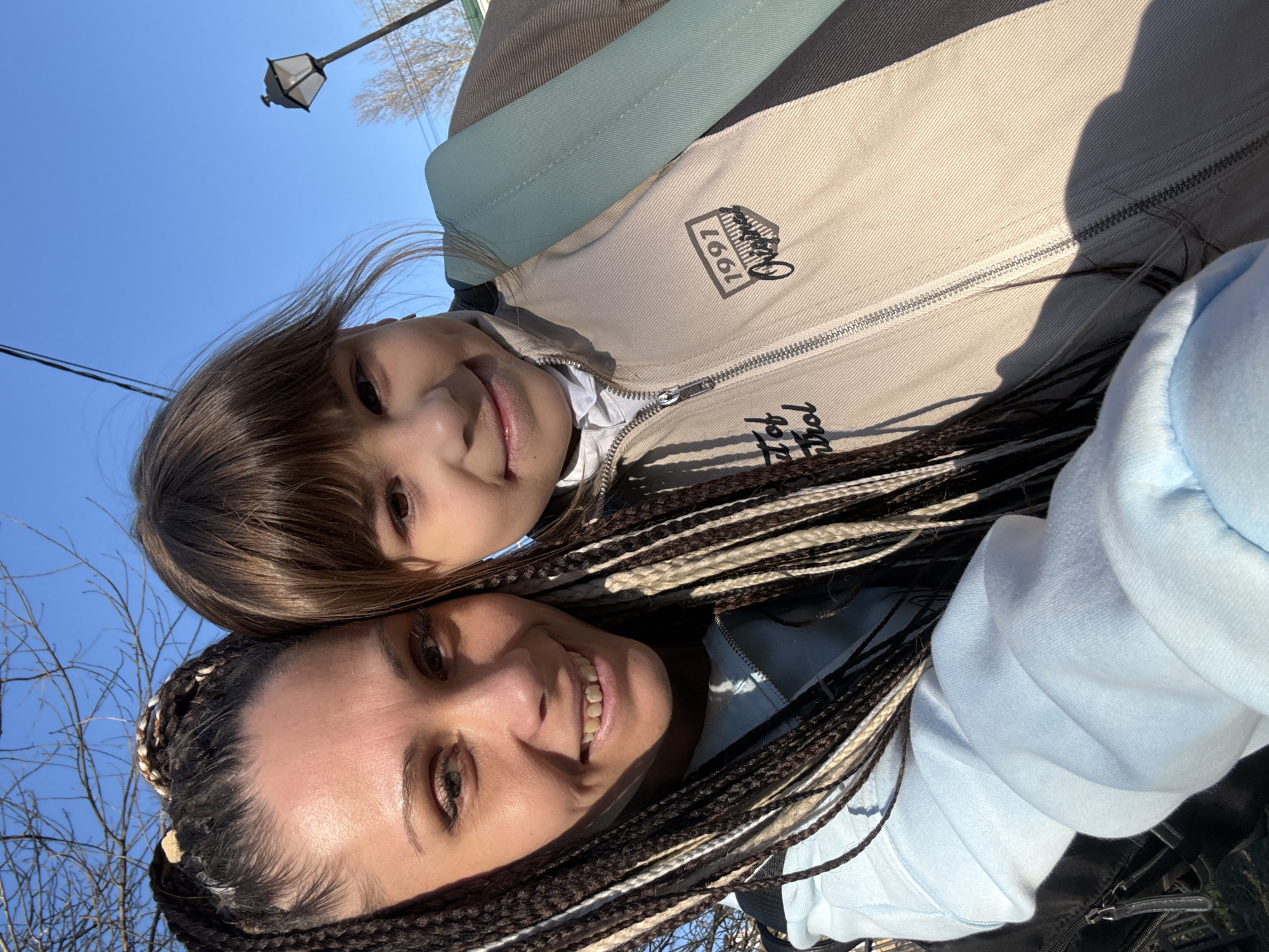 This outdoor selfie, taken in Rusanovo, Russia, captures two smiling females: an adult and a child. The adult, in the foreground, has dark skin and long, elaborate dark and light-colored box braids. She wears a light blue hooded sweatshirt and smiles broadly. The child, positioned above and behind the adult, has fair skin and light brown hair with bangs. She is dressed in a multi-paneled jacket with beige, dark grey, and light blue sections, over a white undershirt. On the beige part of her jacket, a patch clearly displays "1991" and partially visible stylized text that includes "Control." Both individuals are smiling, conveying happiness and contentment. The setting is outdoors under a clear, bright blue sky, indicating a sunny daytime scene. Direct sunlight casts visible shadows on their clothing. In the upper background, a street lamp and bare tree branches are discernible against the sky.