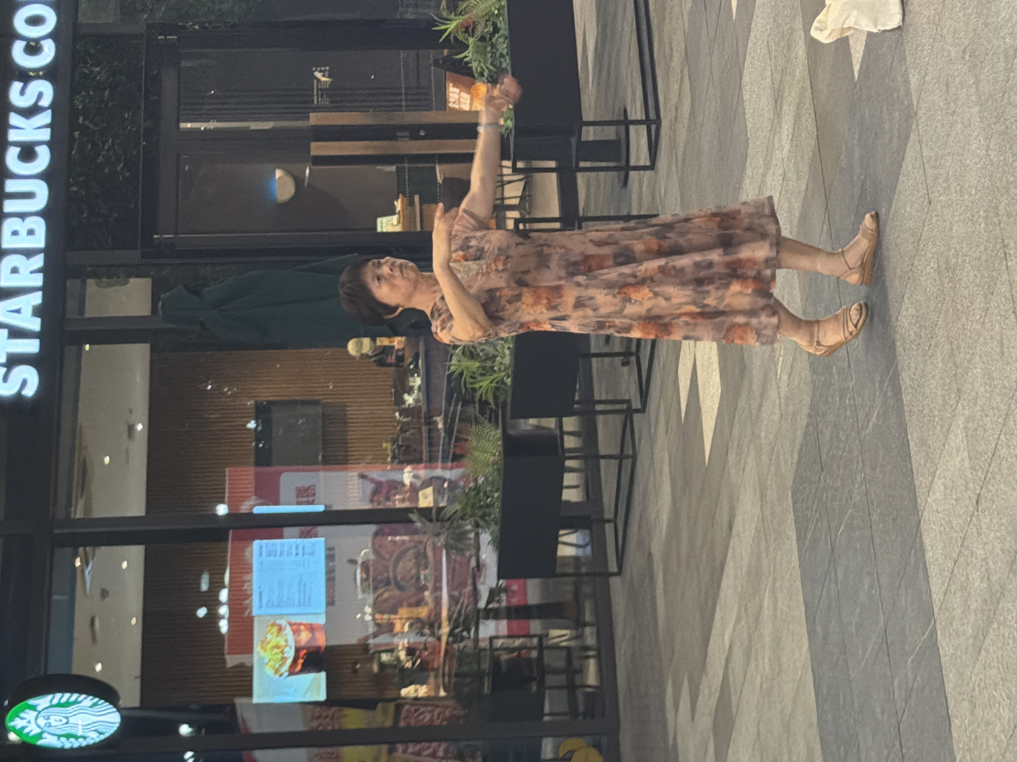 An East Asian woman in a floral dress is performing a slow-motion movement with her arms outstretched in front of a Starbucks. The woman is positioned in the center of the frame, and her body is angled to the right. She has short, dark hair and is wearing light-colored sandals. The background shows the exterior of a Starbucks with a large illuminated sign. In front of the Starbucks, there are several planters with green foliage and some tables and chairs. The ground is paved with grey tiles. The lighting suggests it is daytime, with some shadows cast on the ground. The image appears to be taken in a public outdoor space, likely a pedestrian area in the city of Jiangjin, China.