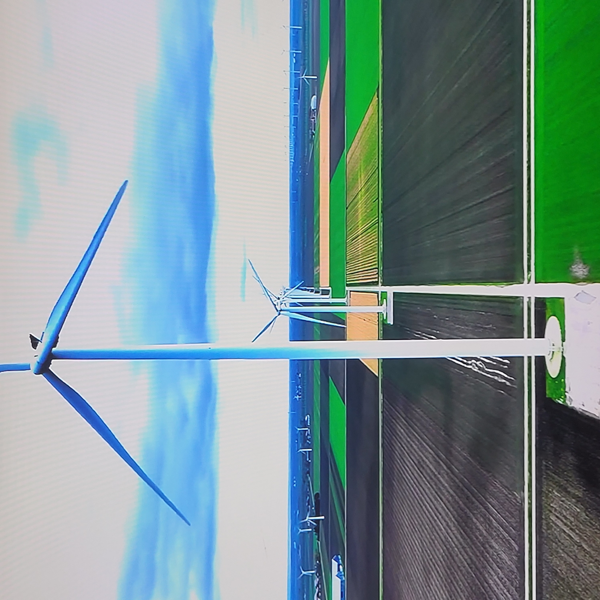 The image depicts a wind farm situated amidst agricultural fields. The turbines are aligned in rows, with the blades extending into the sky. The surrounding landscape consists of alternating patches of green and dark fields, possibly indicating different crops or stages of cultivation. The sky is partly cloudy. There is no indication of the location being Yerevan, Armenia. There are no people visible in the frame.